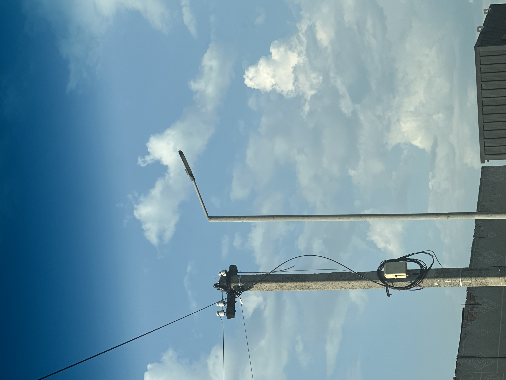The image shows a concrete utility pole with electrical insulators and wires against a blue sky with scattered white clouds. A street lamp is visible to the right of the pole. A small, light-colored electrical box is attached to the utility pole, with coiled black cables around it. In the background, a low, weathered wall with a patterned design and some exposed bricks is visible, along with a dark-colored, corrugated metal structure. The scene appears to be outdoors in an urban or semi-urban setting, possibly in Shatava, Ukraine, given the context provided. There are no people or animals in the image.