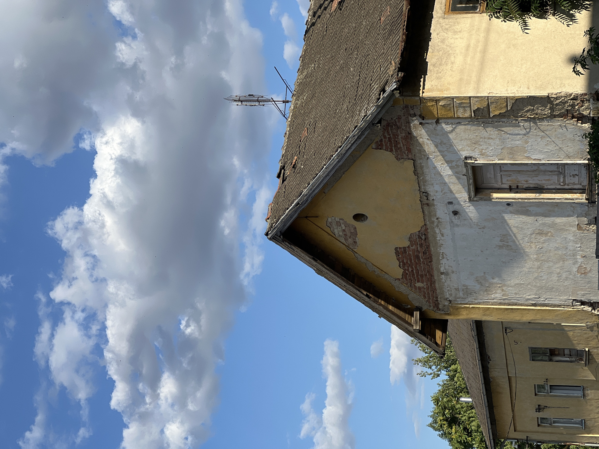 The image shows the exterior of a dilapidated building, possibly a house, in Újezd u Brna, Czechia. The building has a weathered, yellow facade with sections of exposed brick and crumbling plaster. A rustic, light-colored wooden door is visible on the front of the structure. The roof is covered with corrugated material and appears to be in disrepair, with some tiles missing. A tall antenna, likely for telecommunications, is mounted on the roof. The sky above is blue with scattered white clouds. Lush green trees can be seen in the background. The overall impression is one of neglect and age. There are no people visible in the image.