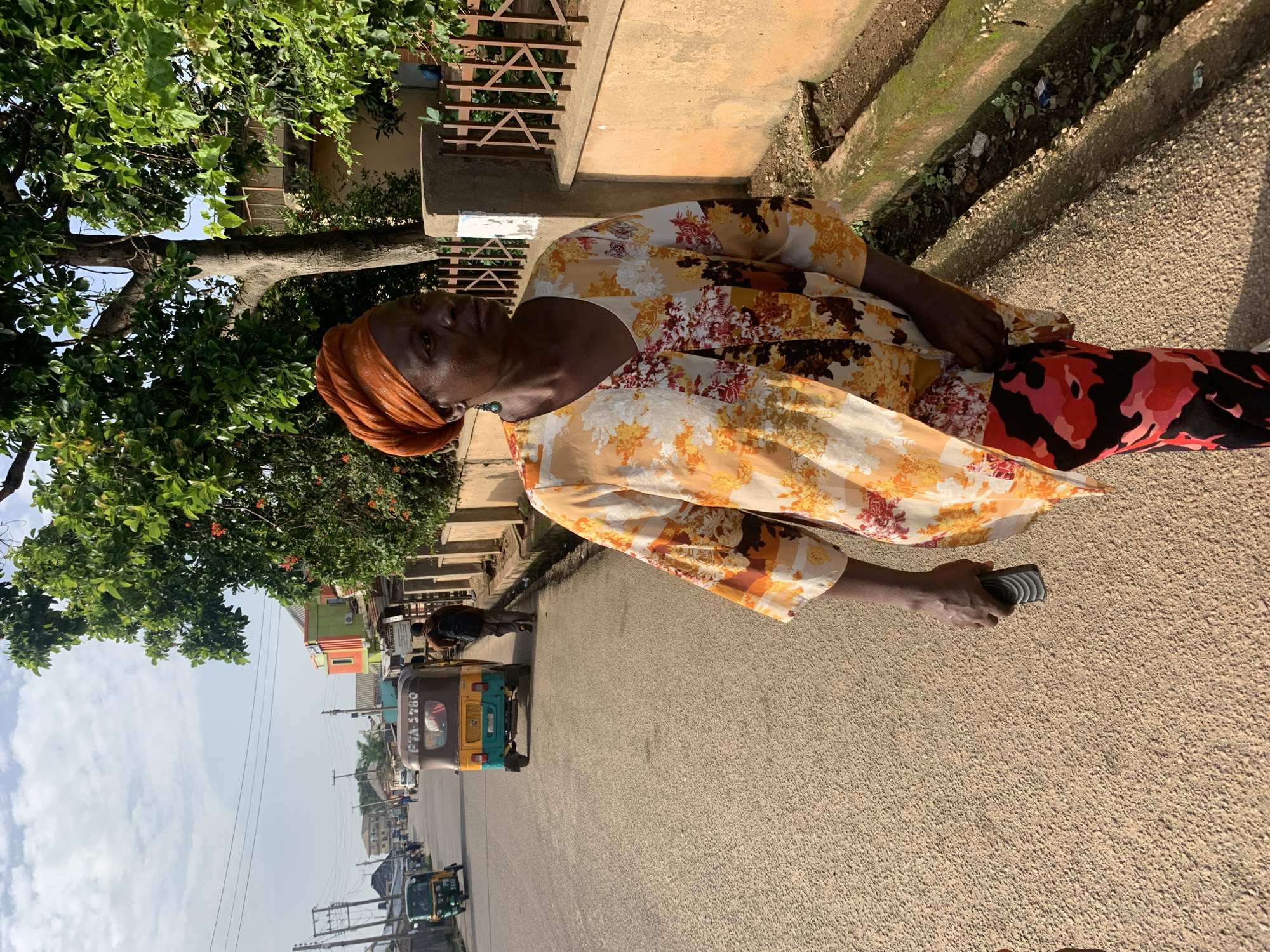 This is a vertical, outdoor, eye-level medium shot of a woman standing on a paved street in Jos, Nigeria. The woman is wearing a floral print dress with a yellow, orange, and white pattern, and an orange headscarf. She has dark skin and is looking towards the camera. Her right hand is holding a grey object.
The scene appears to be a busy street, with several auto-rickshaws (also known as Keke Napeps) visible in the background. Some buildings and trees line the street. The lighting suggests it is daytime, likely late morning or early afternoon, with clear skies and sunny weather.
The woman is the main subject, and her pose and gaze suggest she is aware of being photographed. The presence of the auto-rickshaws indicates an urban or semi-urban environment. There is no discernible text in the image.