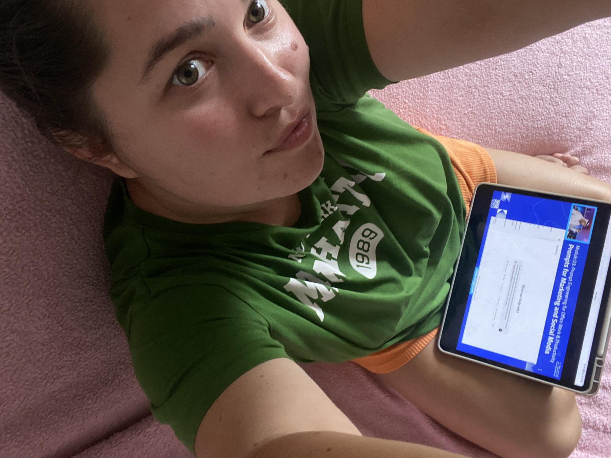 A young woman with brown hair and green eyes is lying down on a pink textured surface. She is wearing a green t-shirt with "MAHAKAN 1989" printed in white lettering and orange shorts. Her left arm is bent and resting on her lap, where a tablet is positioned. The tablet screen displays content related to "Prompts for Marketing and Social Media" and "Module Q3: Prompt Engineering for Office Work & Productivity." Her gaze is directed towards the camera. The scene appears to be indoors, in a domestic setting. No specific location in the city of Perm, Russia, can be determined from the provided image.