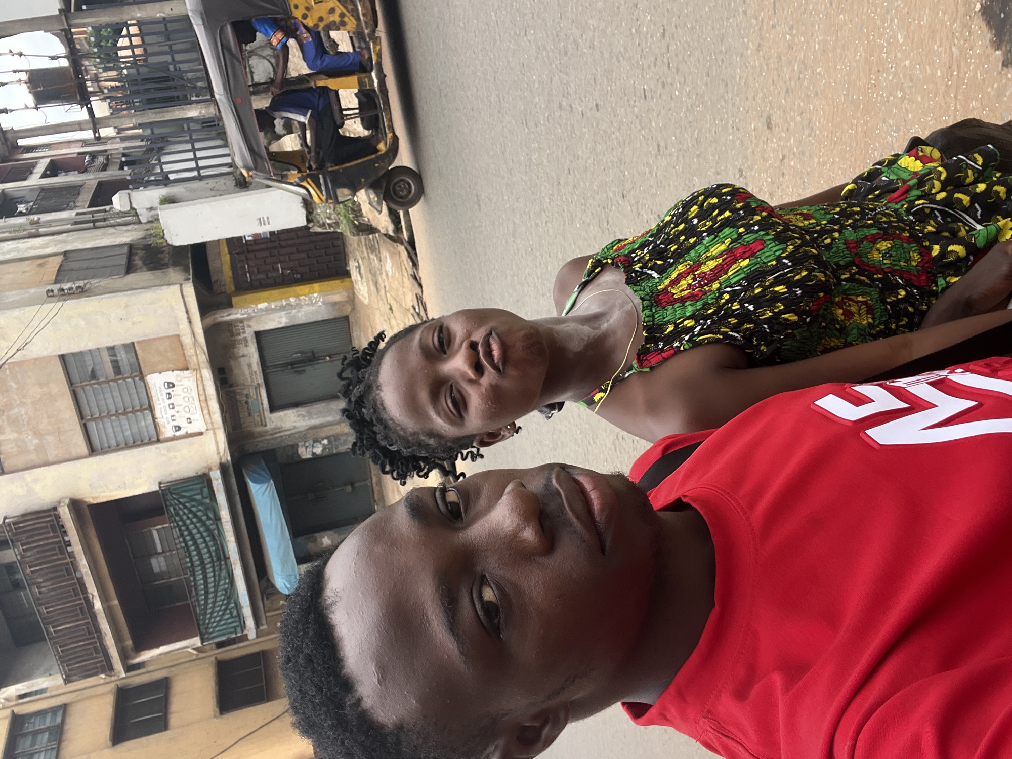 In Nkpor, Nigeria, a young Black man in a red shirt with white lettering and a young Black woman in a colorful patterned dress stand on a paved street. The man is closer to the camera, looking slightly to his left. The woman is to his right, facing the camera with her lips pursed. Behind them, a yellow and black auto rickshaw, also known as a "Keke Napep," is driving on the street. Two men are visible inside the auto rickshaw; one appears to be driving, and the other is seated behind him. The background features a multi-story building with balconies and windows, typical of urban architecture in Nigeria. The sky is overcast.