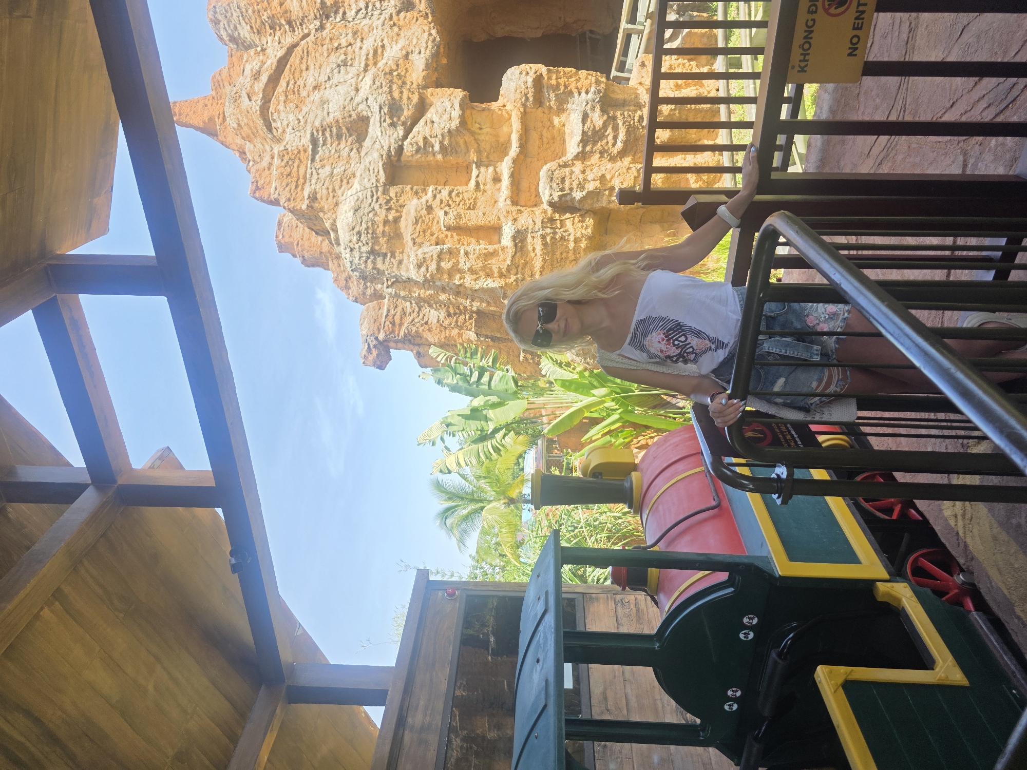 The image captures a woman with blonde hair wearing sunglasses, leaning against a metal railing on what appears to be a colorful, toy-like train. The train, with red, green, and yellow components, stands against a backdrop of tropical foliage including palm trees. Behind the woman, there is an artificially-made, light-brown, rocky structure that resembles a cave. A yellow sign that reads "KHÔNG ĐI - NO ENT" is affixed to the railing. The scene is sunlit, suggesting daytime. The setting evokes a recreational or amusement park environment. The location is Đà Nẵng, Vietnam.
