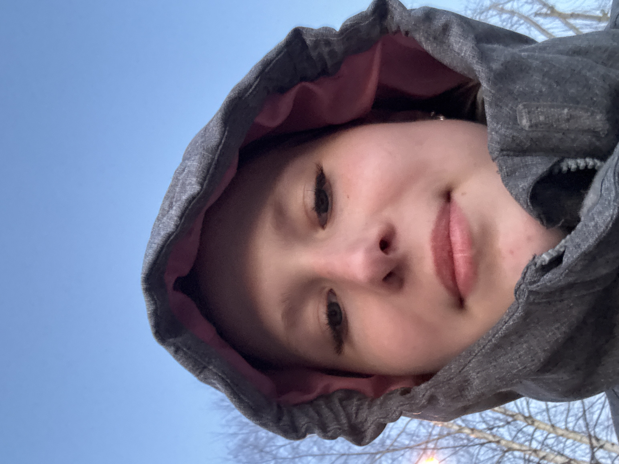A young person with fair skin and light-colored eyes is wearing a gray hooded sweatshirt with a pink lining. The hood is up, framing their face. The person is looking down at the camera. The background consists of a blue sky and bare branches of trees. The image appears to have been taken outside in Berezniki, Russia, based on the sky and trees.