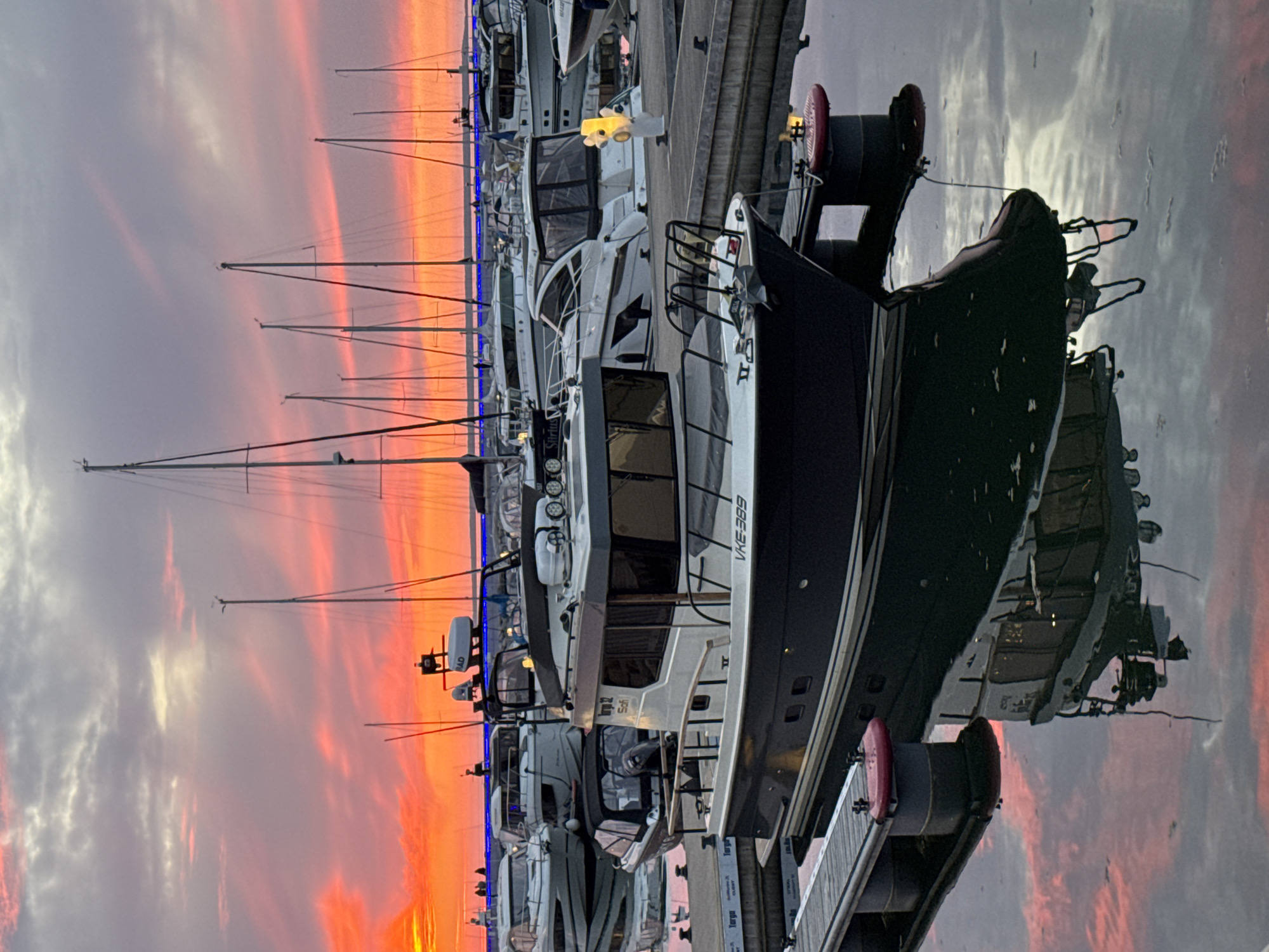 The image depicts a serene marina scene in Tallinn, Estonia, at either sunset or sunrise, evidenced by the dramatically colored sky. The original image is oriented vertically, but the scene shows horizontally docked boats.
In the foreground, a prominent dark-hulled motor yacht with a white superstructure is moored at a pier. The registration "VHE-389" is clearly visible on its side. Its reflection, along with the vibrant sky, is visible in the calm water. Along the piers, several other motor yachts, predominantly white, are docked. One of these boats has "SIRIUS" visible on its side, and another displays a "SIMRAD" radar dome. The slender masts of numerous sailboats are silhouetted against the bright sky, indicating a diverse collection of vessels in the harbor.
The sky is a spectacular blend of intense orange, red, pink, and purple hues, particularly concentrated near the horizon, gradually transitioning to lighter grey and blue tones higher up. Clouds are scattered across the sky, catching and reflecting the vivid light. Artificial blue lighting emanates from some of the boats, and a distinct yellow decorative light fixture is visible on one of the white yachts. Red-capped bollards or pedestals line the docks, one of which is illuminated and bears the text "Torgu". The tranquil water mirrors the striking sky and the outlines of the boats and docks. No people or specific activities are visible, emphasizing the peaceful and picturesque ambiance of the marina at dawn or dusk.