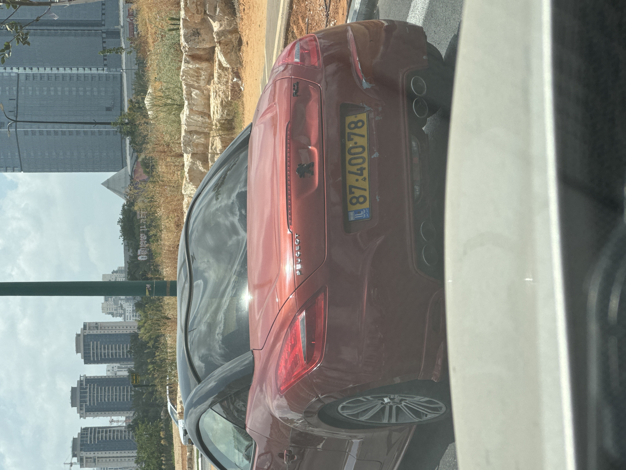The image captures the rear of a red Peugeot RCZ sports car driving on a street in Petah Tikva, Israel. The car has a black retractable hardtop and dual exhaust pipes. Its license plate reads "87-400-78". The car appears to have some damage on its rear bumper and right taillight. In the background, several modern high-rise buildings are visible, along with dry vegetation and some construction. The sky is cloudy.