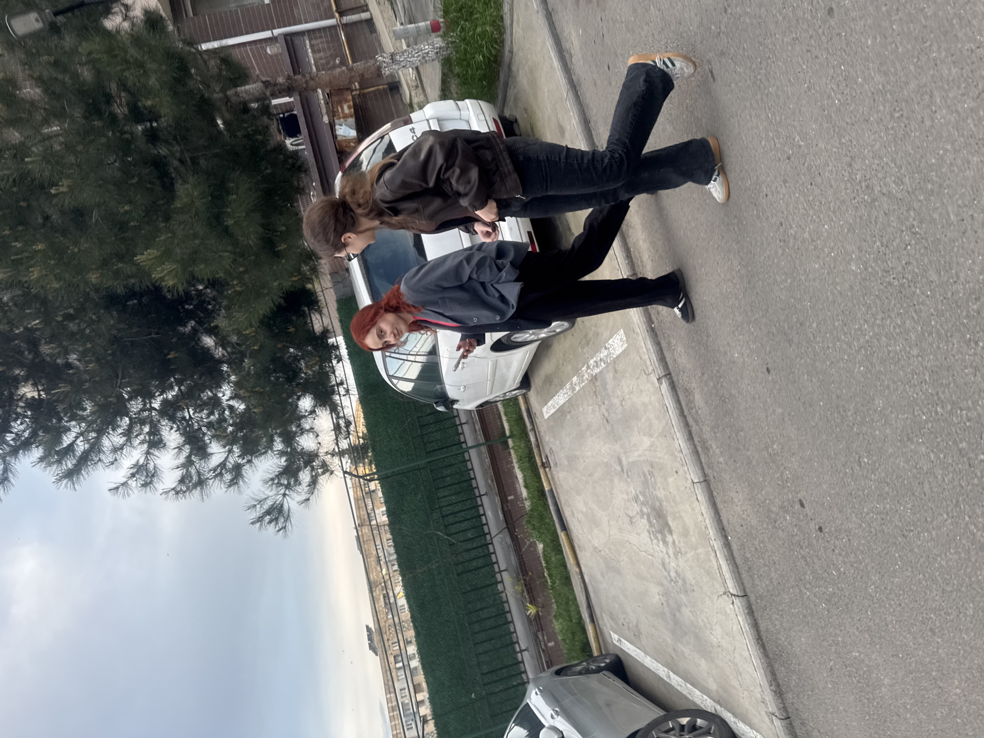 Two women stand on a paved surface, likely a parking lot or road, next to parked cars and a green fence. One woman has auburn hair and wears a gray jacket and black pants. The second woman has brown hair and wears a brown jacket and black pants. The background includes trees and buildings, and it appears to be daytime. The image was taken in Tbilisi, Georgia.