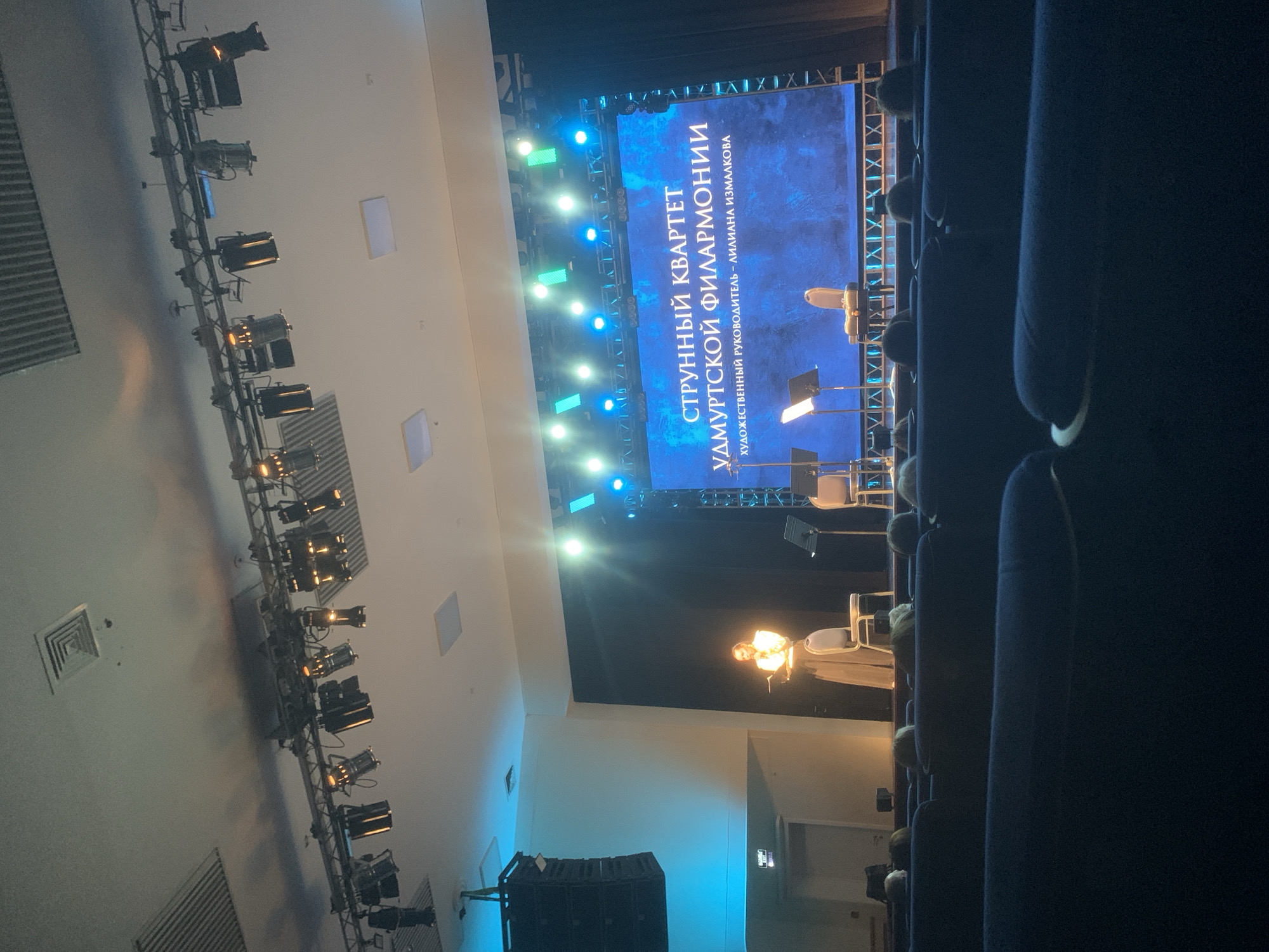 The image depicts the interior of a concert hall in Izhevsk, Russia, likely the Udmurt State Philharmonic. A string quartet is performing on stage, visible below a large screen displaying text in Russian: "СТРУННЫЙ КВАРТЕТ УДМУРТСКОЙ ФИЛАРМОНИИ" and "ХУДОЖЕСТВЕННЫЙ РУКОВОДИТЕЛЬ - ЛИЛИАНА ИЗМАЛКОВА" (String Quartet of the Udmurt Philharmonic, Artistic Director - Liliana Izmalkova). Stage lighting is visible above the performers and the screen. Rows of unoccupied seats face the stage. A person is visible on stage, presumably a performer.