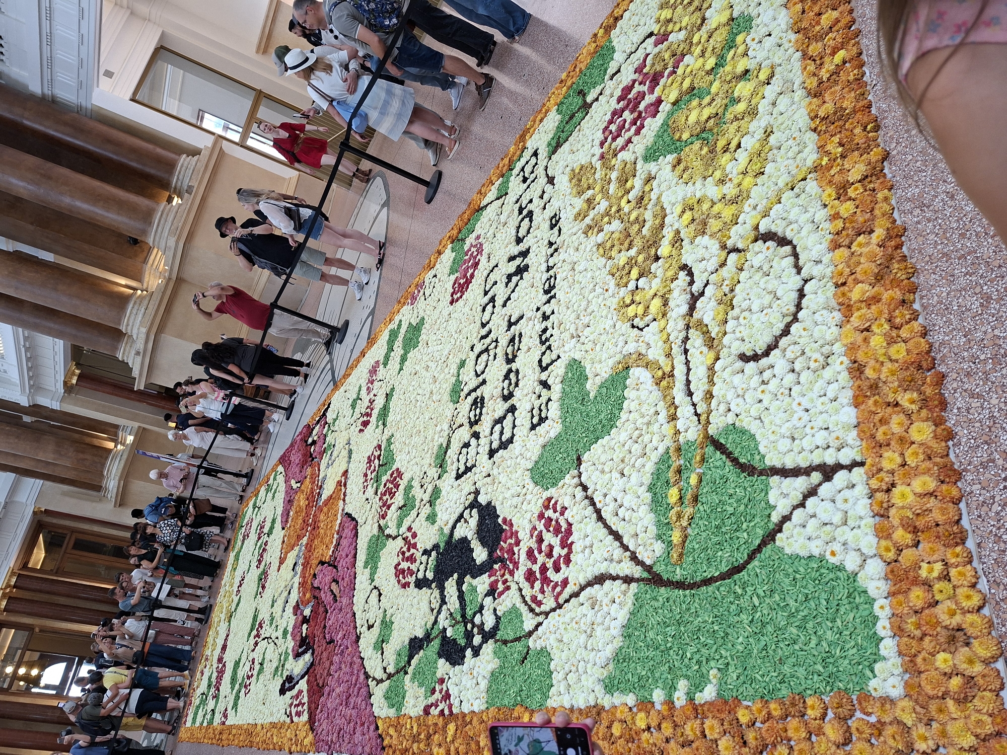The image depicts a large flower carpet, likely located in Brussels, Belgium, given the context of flower carpets often being displayed in the city, most notably at the Grand Place. The carpet features a vibrant display of flowers arranged to form intricate patterns and text. The text visible on the carpet reads "Belgian Beer World Experience".
Numerous people are standing around the perimeter of the flower carpet, observing and taking photographs. They are dressed in casual clothing, suggesting a public event or attraction. The setting appears to be an indoor public space, possibly a museum or exhibition hall, indicated by the high ceilings, architectural columns, and flooring. Some individuals are holding up phones to capture images of the display. The overall atmosphere seems to be one of admiration and engagement with the floral artwork.
