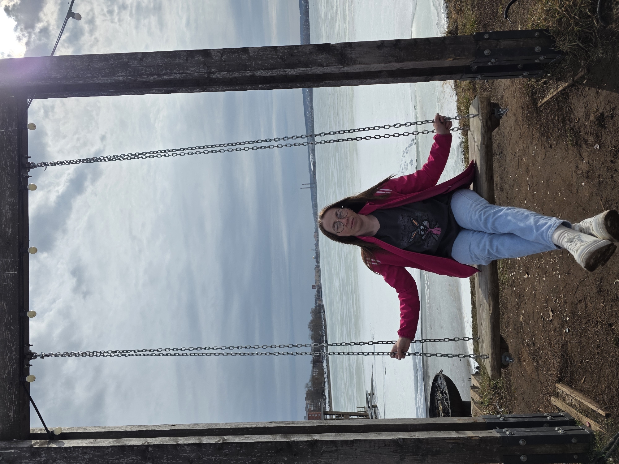 A young woman is sitting on a swing set near a body of water covered in ice, likely in the city of Votkinsk, Russia. She is wearing glasses, a t-shirt, a pink jacket, blue jeans, and white sneakers. The swing set is made of wood and metal chains. The sky is cloudy. The ground around the swing set is dirt and grass. In the distance, a city skyline is visible across the icy water.