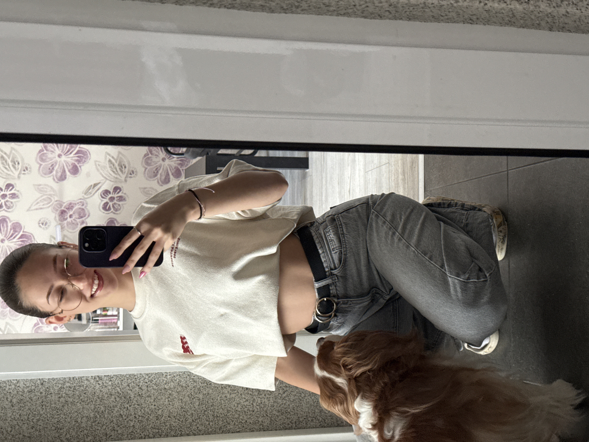 A young Caucasian woman with dark hair pulled back and wearing glasses is taking a mirror selfie. She is wearing a cream-colored cropped t-shirt, light grey denim jeans, and a black belt with a double ring buckle. Her left arm is extended, holding a dark-colored smartphone. A small, light brown and white Cavalier King Charles Spaniel dog is partially visible to her left, looking up at her. The background shows a wall with floral wallpaper and a mirror reflecting the scene. The flooring appears to be dark grey tiles. The environment suggests an indoor setting, possibly a residential bathroom or dressing area. There are no specific visual cues to place this image in Sofia, Bulgaria.