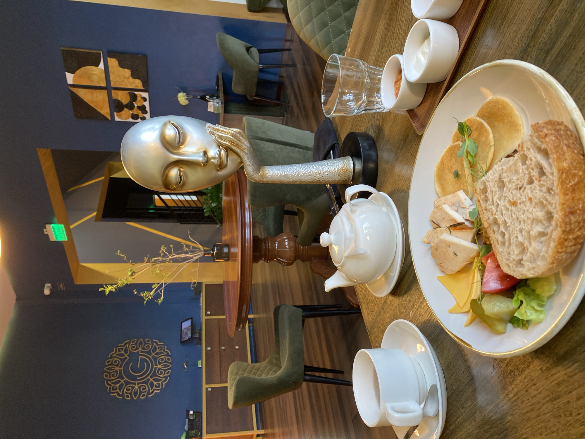 A table in a cafe in Yekaterinburg, Russia, is set with a plate of food including pancakes, bread, sliced chicken, cheese, tomato, and pickles, alongside a teapot, teacup, and sauces. A decorative statue of a head and hand rests on the table near the plate. The cafe interior features a blue wall with gold accents and abstract artwork, olive-green chairs, and decorative plants.