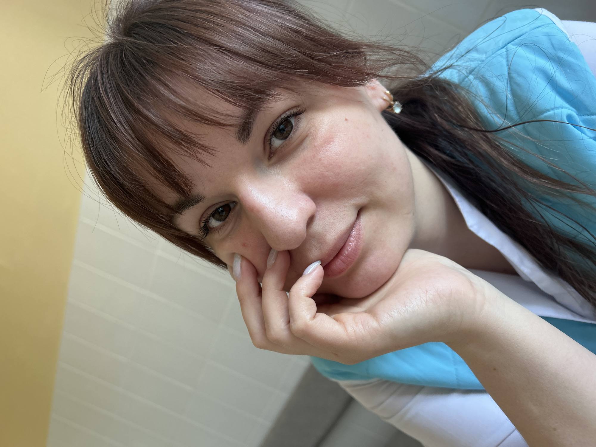 A close-up portrait shows a female individual with medium brown hair, including bangs, and brown eyes. Her complexion is fair, with slight redness on her cheeks and a small blemish on her left cheek. She wears a white garment under a bright blue vest or uniform top. Small gold-colored hoop earrings with light-colored, possibly gemstone, drops are visible. Her left hand, with manicured nails featuring a light, pearlescent polish, rests against her chin and lower left cheek. She looks directly at the camera with a subtle expression. The indoor background displays a light-colored, vertically-patterned wall on the left, with a dark horizontal line segment visible. The upper right background is a solid light yellow. Based on the provided context, the scene is situated in the city of Фоки, Russia.