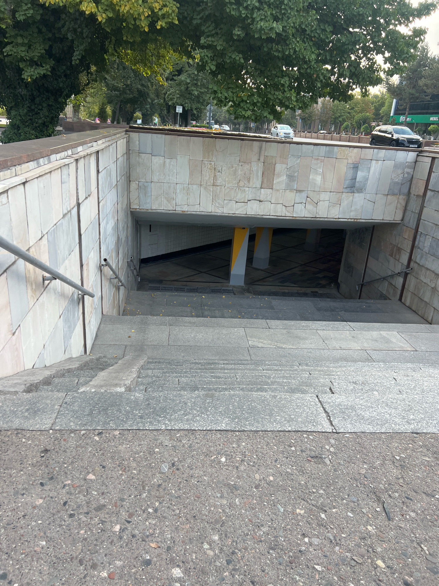 The image shows a view looking down a wide set of outdoor stairs leading to an underground walkway. The stairs are made of a grey, speckled material, possibly granite or concrete. The walls of the staircase and the surrounding area are clad in light-colored, rectangular stone tiles. A metal handrail runs along the left side of the stairs. The entrance to the underground walkway is visible at the bottom of the stairs, with patterned flooring and yellow and grey pillars. Above the underground entrance, a street is visible with trees lining the sidewalk and cars driving on the road. A double-decker bus with green signage is also visible on the street. The overall scene appears to be an urban environment.