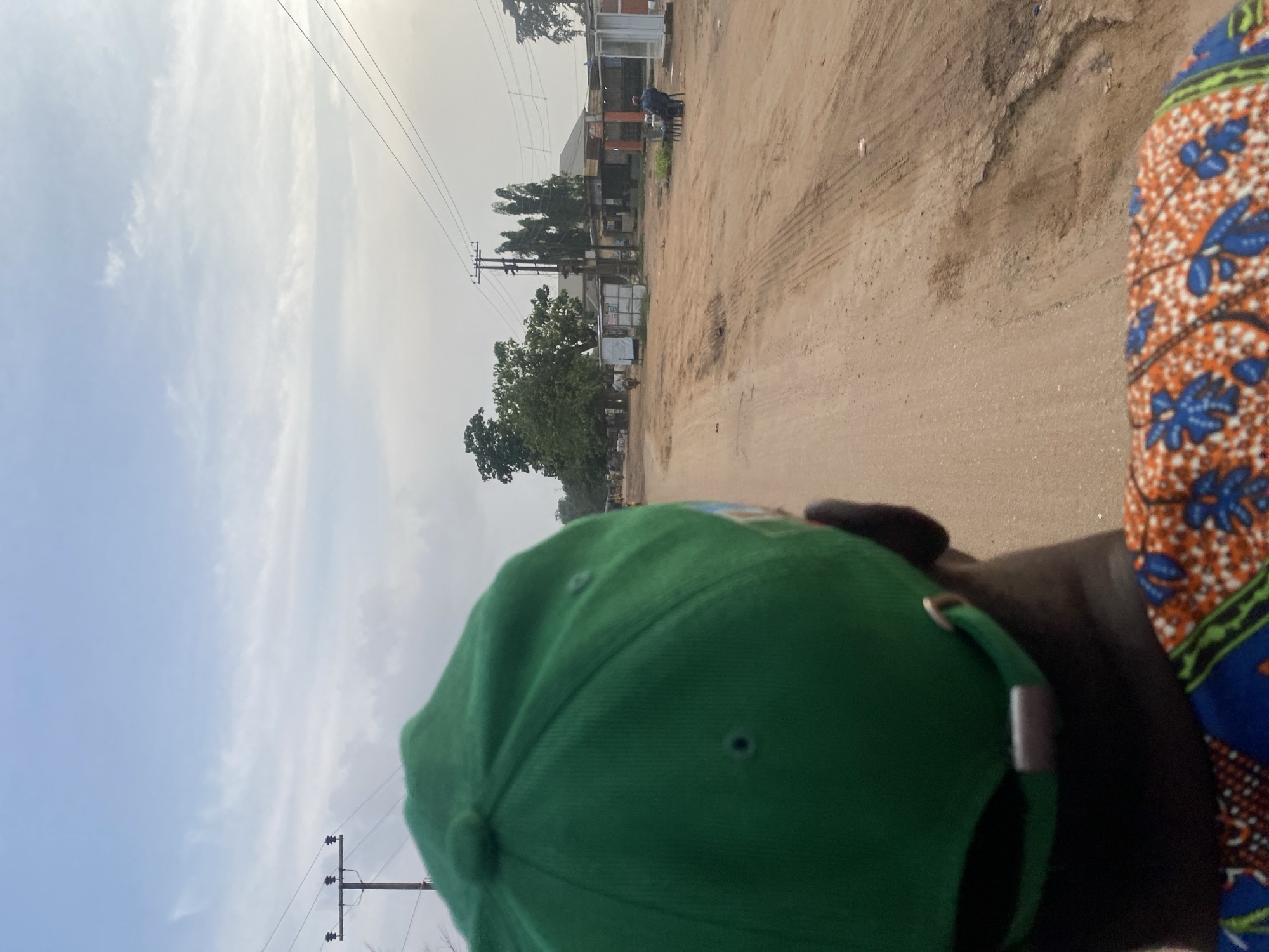 The image captures a close-up view from behind a person wearing a green baseball cap and a vibrant, patterned shirt, looking out at a dusty, unpaved road in Ikorodu, Nigeria. The road appears worn, with tire tracks and some potholes, suggesting it is a common thoroughfare. On either side of the road are scattered buildings, some appearing to be small shops or stalls, with corrugated metal roofs and open fronts. Utility poles with electrical wires run along the side of the road, indicating the presence of electricity infrastructure. In the distance, there are trees and a few people, one of whom is seated at a table outside one of the structures. The sky is partly cloudy, with a bright, overcast appearance suggesting it is daytime, likely in the morning or late afternoon due to the soft light. The overall scene depicts a typical street view in a less developed urban or semi-urban area.