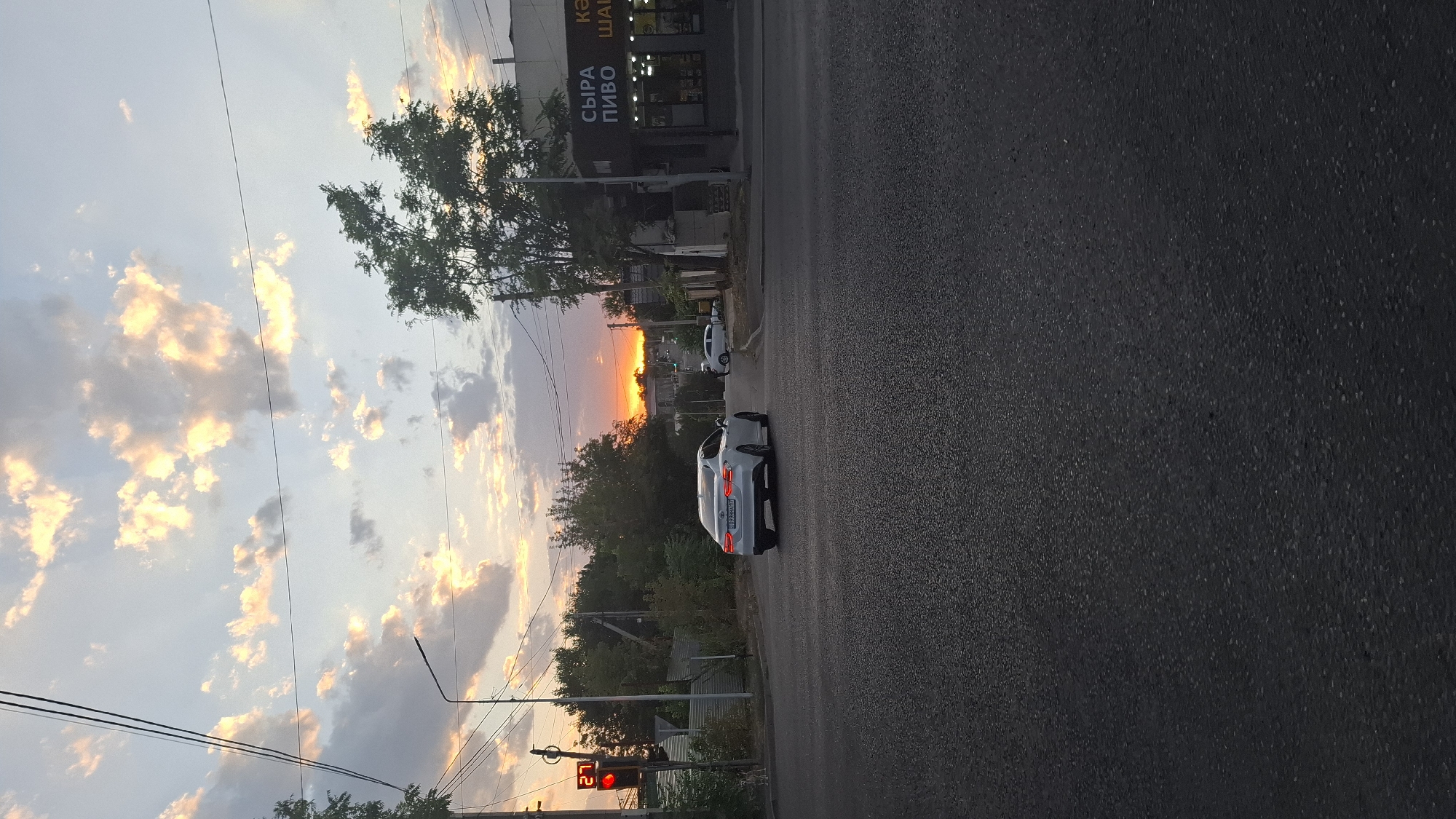 The image depicts a street scene at sunset in Shymkent, Kazakhstan. A white sedan is captured in motion, turning at an intersection. Traffic lights are visible, with one showing a red light and the number "27" displayed. Buildings line the street, one of which features signage in Cyrillic that translates to "Cheese Beer Shawarma." Trees and power lines are present in the background, contributing to an urban landscape. The sky is filled with dramatic clouds illuminated by the setting sun, casting a warm glow over the scene. The road surface is asphalt.