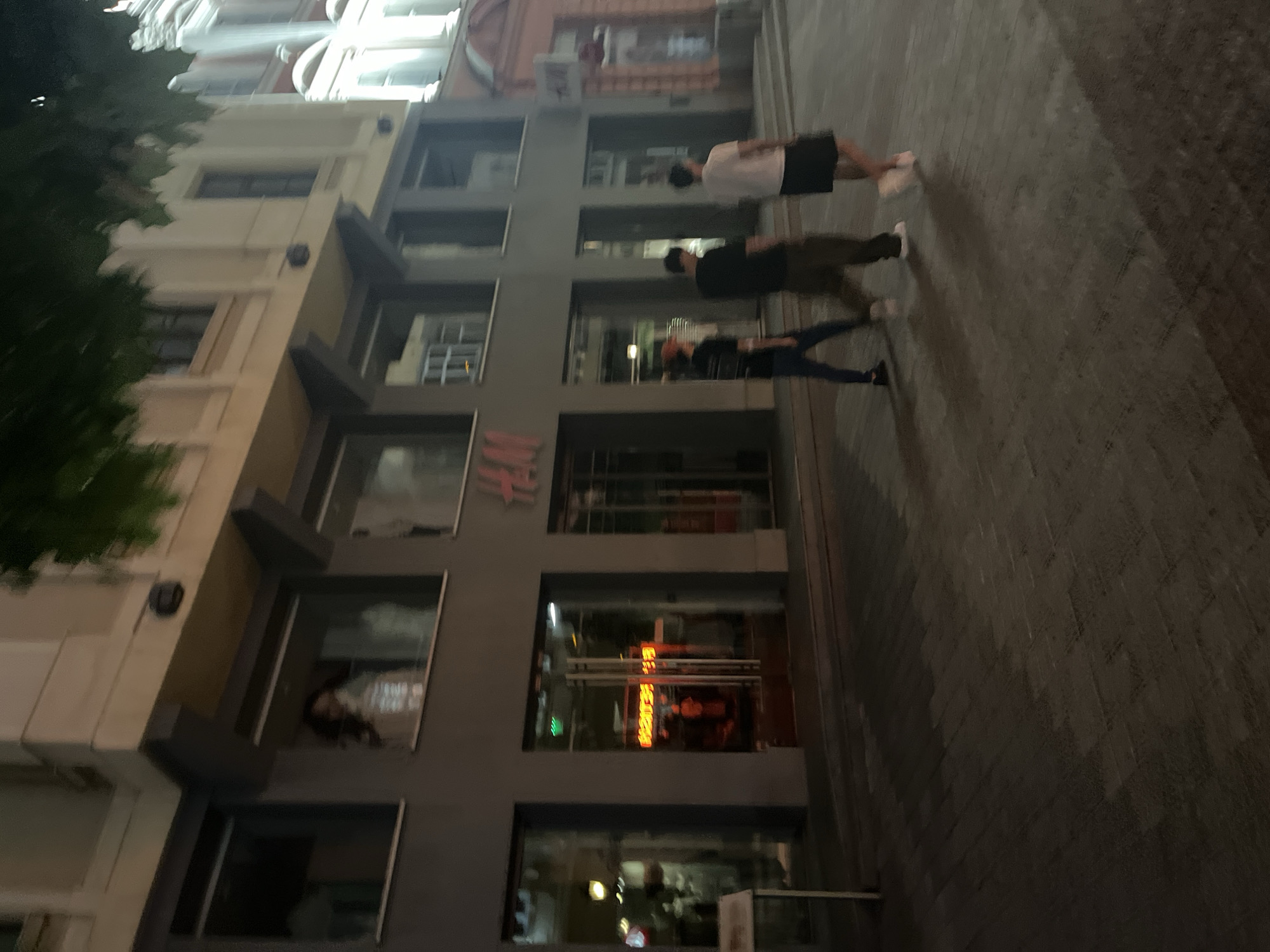 This nighttime image depicts Knyaz Alexander I Street in Plovdiv, Bulgaria. The central focus is a multi-story building featuring an H&M retail store, identifiable by prominent red "H&M" logos on its grey facade. On the second floor, a mannequin is visible through a large window. To the immediate right of the H&M entrance, through a double glass door, a reversed neon sign displaying "FETISH" is illuminated. Further to the right, a distinctly ornate, light-colored building with multiple windows and architectural details is visible. Three male individuals are captured walking from left to right across the paved pedestrian area in the foreground. The individual on the left wears a dark t-shirt, dark pants, and a light cap. The middle individual wears a dark t-shirt and dark pants. The individual on the right wears a white t-shirt, dark shorts, and light-colored shoes. The ground consists of patterned paving stones. Overhead, blurred dark foliage suggests trees. The scene is illuminated by artificial street and building lights.
