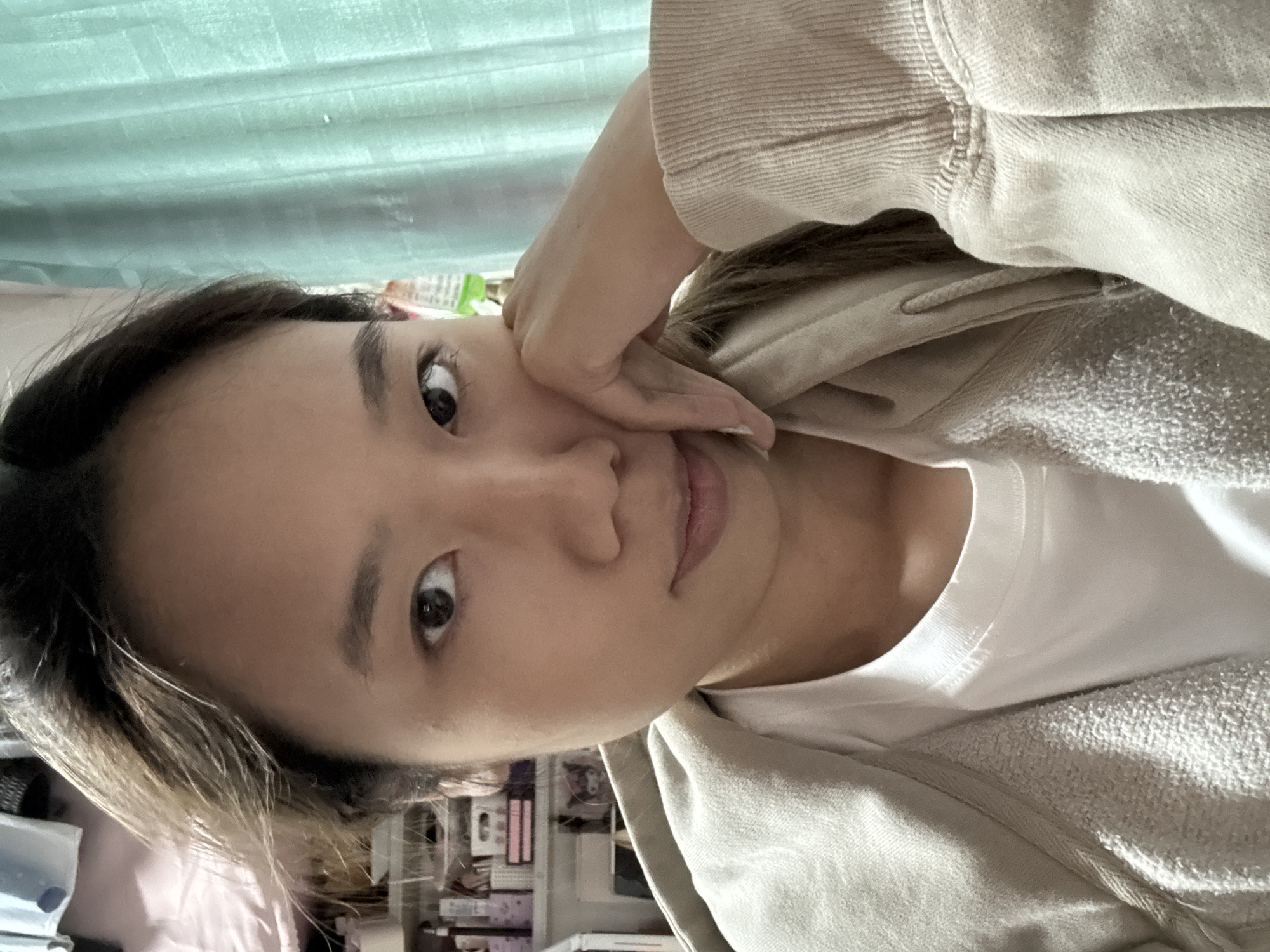 A young adult female faces the camera at an angle, her right hand resting on her cheek. She has dark hair and eyes, and is wearing a white t-shirt under a beige hooded sweatshirt. Behind her is a light green curtain, and in the lower left corner, a shelf with various objects is visible. The location is suggested to be Nan'an District, China.