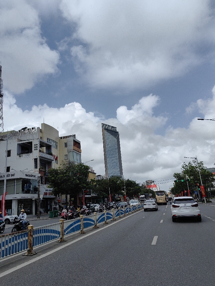 The image captures a daytime street scene in Thuận Hóa Ward, Vietnam, under a sky with prominent white and grey cumulus clouds and patches of blue. The viewpoint is from a low angle on an asphalt road featuring white lane markings. A blue and white patterned metal barrier, showing some rust, delineates the left side of the road, separating the main vehicular lanes from an area with parked motorcycles and a sidewalk.
Along the left side, a row of multi-story buildings with varied architectural styles line the street. These include commercial establishments on the ground floor, indicated by Vietnamese signage such as "NHÀ XUẤT DƯ" and "ANH KHÁNH". Green-leafed trees are planted along this stretch. A tall, slender, glass-clad skyscraper with a distinct, possibly trapezoidal, shape dominates the mid-background, featuring a logo near its apex. To the far left, a metal lattice tower is partially visible.
Traffic on the road moves away from the viewer. A white SUV with a visible license plate segment '75A-XXX.XX' occupies the right lane in the foreground. A silver sedan is positioned in the left lane ahead of the SUV. Further down the street, a light-colored bus and additional vehicles are visible. Numerous motorcycles are present along the left side of the road, behind the barrier, with several individuals wearing helmets. One motorcyclist in a white helmet and light-colored jacket is depicted in the immediate foreground, facing left. Red and white banners are affixed to poles along the street.