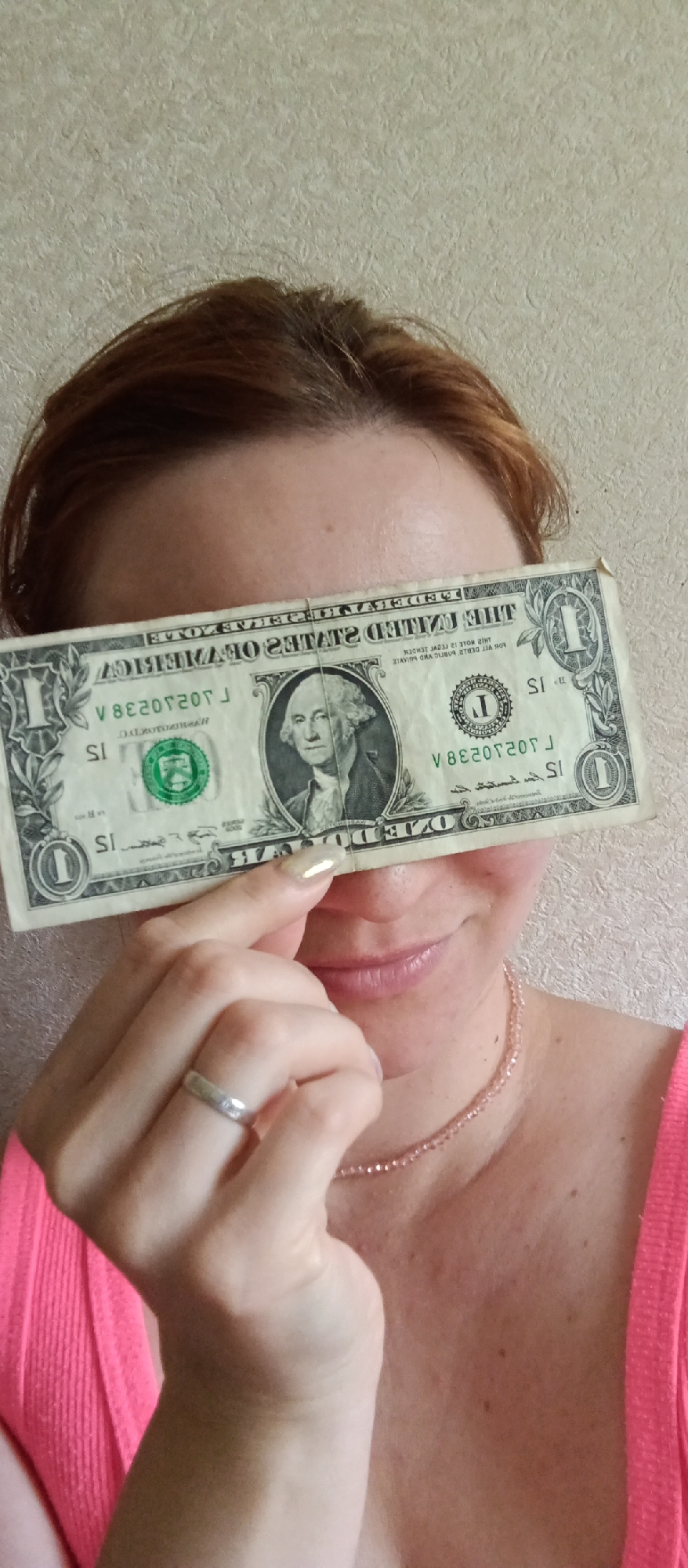 A woman is holding a one-dollar bill over her eyes. She is wearing a silver ring on her left ring finger and a pink beaded necklace. She has reddish-brown hair and is wearing a pink top. The background is a beige wall.