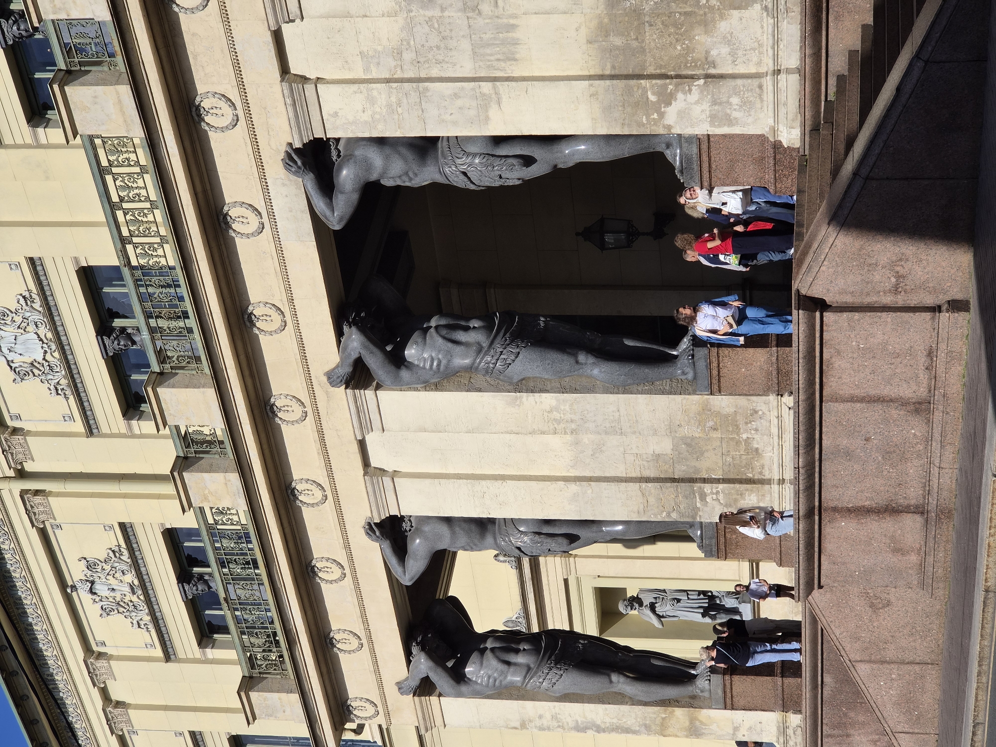The image shows a view of the Atlantes of the New Hermitage Museum, a classical building with a yellow facade and grand columns. Four imposing, muscular statues of men, carved from dark stone, are depicted holding up the entablature with their hands above their heads. The statues are arranged in a line, creating a colonnade. Between the statues, architectural details like windows and balconies with decorative iron railings are visible on the upper floors of the building.
In the foreground, a group of people are gathered on a wide, stepped entrance made of polished pink granite. The people appear to be tourists, dressed in casual clothing. Some are looking at the statues, others are talking amongst themselves or looking at their phones. One woman in the foreground is wearing a white top and blue jeans, holding a phone. Another woman to her left is wearing a dark top and blue jeans. Further to the left, a man in a blue shirt and jeans is standing.
Behind the statues, a statue of a classical figure is visible in an alcove. A lantern is also mounted on the wall behind the statues. The sky is bright and appears to be clear, suggesting a sunny day. The overall impression is a tourist scene in front of a historically significant architectural landmark.