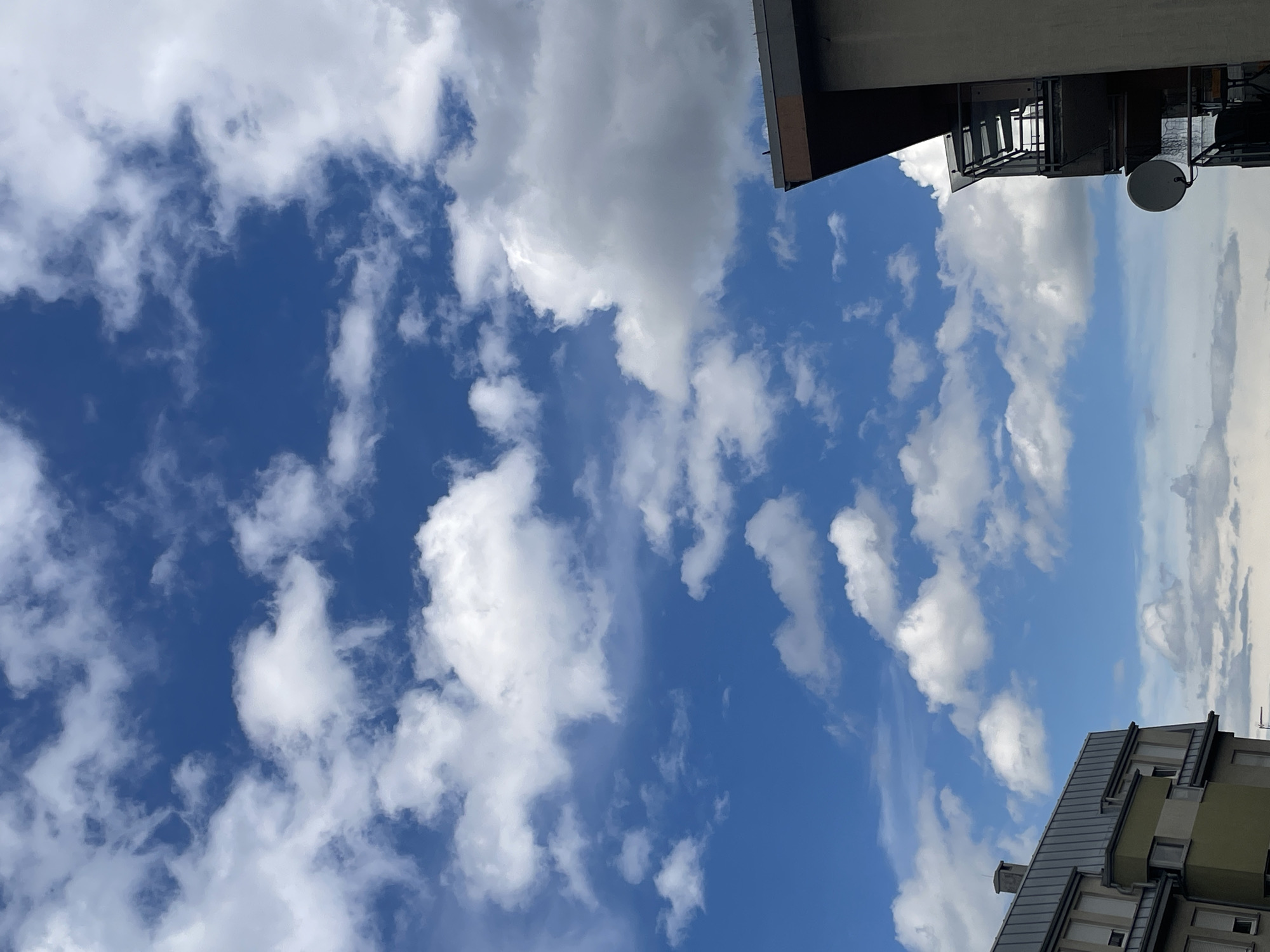 The image shows a view of the sky from what appears to be an urban location. The sky is blue with scattered white clouds. Portions of buildings are visible at the top and bottom edges of the frame. The building at the top appears to have a balcony with a satellite dish. The building at the bottom has a multi-storied facade with windows and a grey roof.
