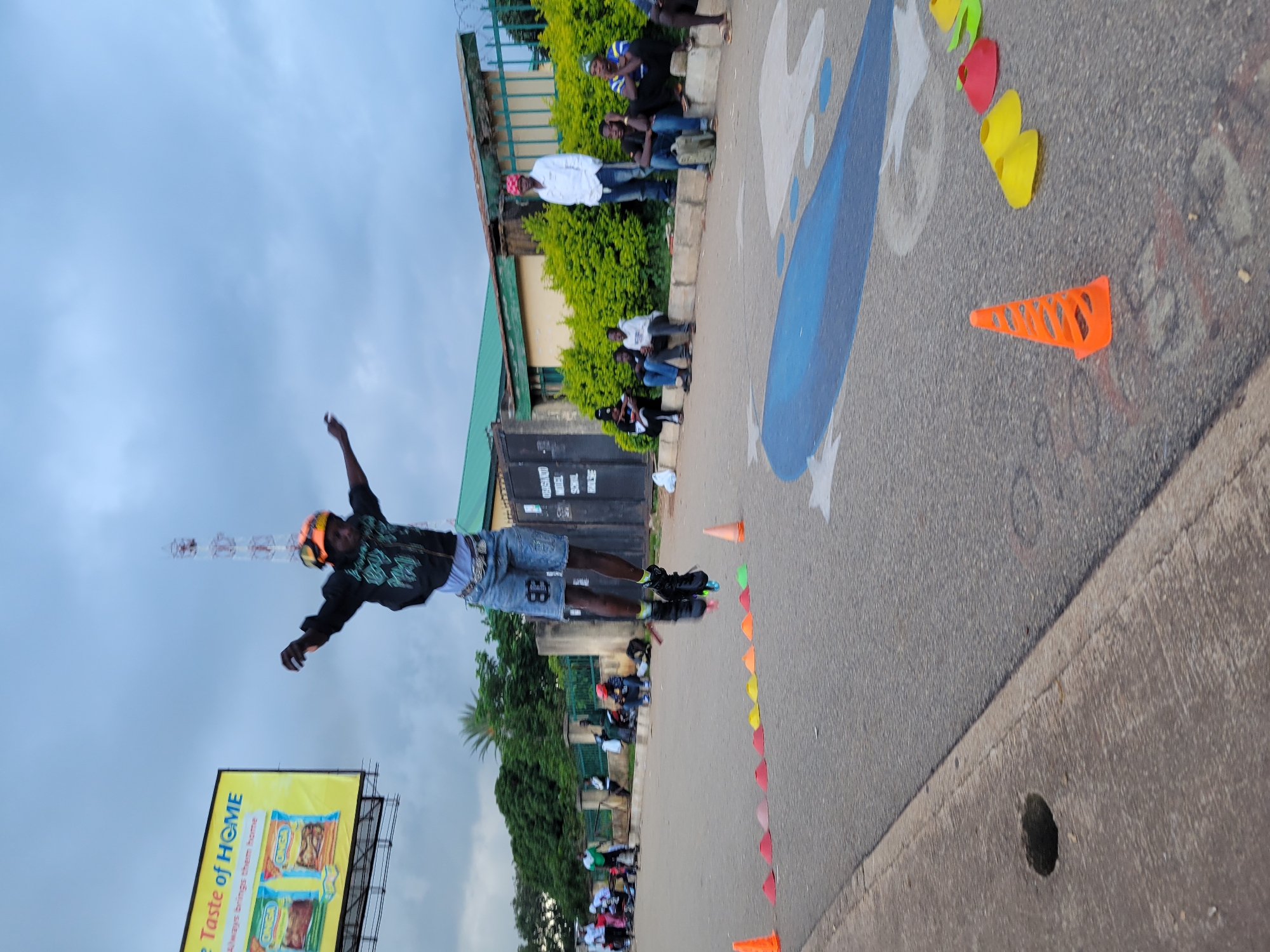 The image captures an outdoor scene on a cloudy day, likely in Jos, Nigeria, given the context. In the foreground, a young person on rollerblades is mid-air, performing a jump. They are wearing a black hooded jacket with a green design, denim shorts with the number "83" visible, and rollerblades. Their arms are outstretched for balance. The ground is paved with asphalt, marked with a large blue painted shape, possibly part of a game or training course. A line of colorful cones, including orange, red, yellow, and green, are placed in sequence on the road. To the left, a concrete sidewalk borders the road, with a circular hole visible.
In the mid-ground, several individuals are seated or standing along a low wall. They appear to be spectators or participants waiting for their turn. A fence separates the road area from a building with a green roof and yellow walls. In the background, trees and a cell phone tower are visible against the overcast sky. Further to the left, a billboard advertises "Taste of HOME" with images of food products. The overall atmosphere suggests a recreational or sporting event taking place in an urban or semi-urban environment.