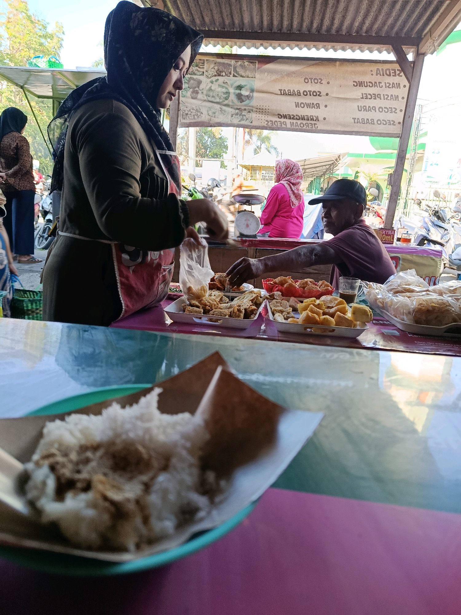 An outdoor food stall in Bululawang, Indonesia, is captured in this medium-shot photograph. The foreground features a close-up of a serving of white rice with toppings on a green plate, set on a table with a glossy, reflective surface that shows a blurred purple and turquoise color palette. Behind the rice, a woman wearing a dark hijab and an apron is visible, attending to food in plastic bags. She is in the process of serving or preparing food. To her right, an elderly man with a dark baseball cap is seated behind a display of fried snacks and dishes, and he is reaching for a glass of coffee. Behind them, a banner listing menu items is displayed. The background reveals more of the market atmosphere, with other stalls, motorbikes, and trees visible under a corrugated metal roof. The scene appears to be a busy street-side food vendor environment.