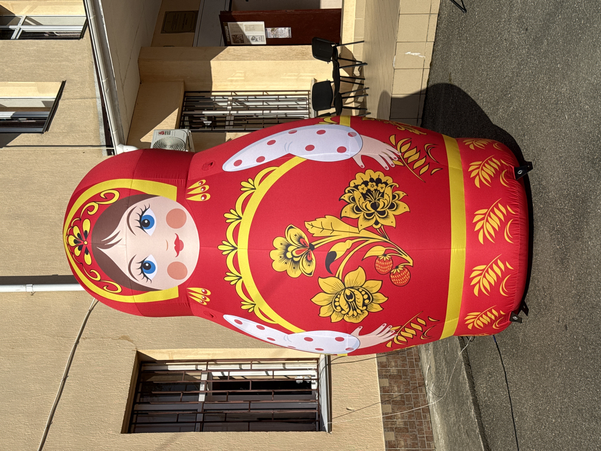An oversized, inflatable matryoshka doll, predominantly red with yellow and white accents, stands inflated on an asphalt surface in front of a light brown, two-story building. The building features barred windows on the ground floor and regular windows on the upper floor, as well as an air conditioning unit and an entrance with a sign indicating "INFORMATION." Two black chairs are placed on a tiled step leading up to the entrance. The matryoshka doll is adorned with a painted face featuring blue eyes, rosy cheeks, and red lips, as well as traditional floral patterns. The scene appears to be outdoors, likely in a public space within the city of Progress, Russia.