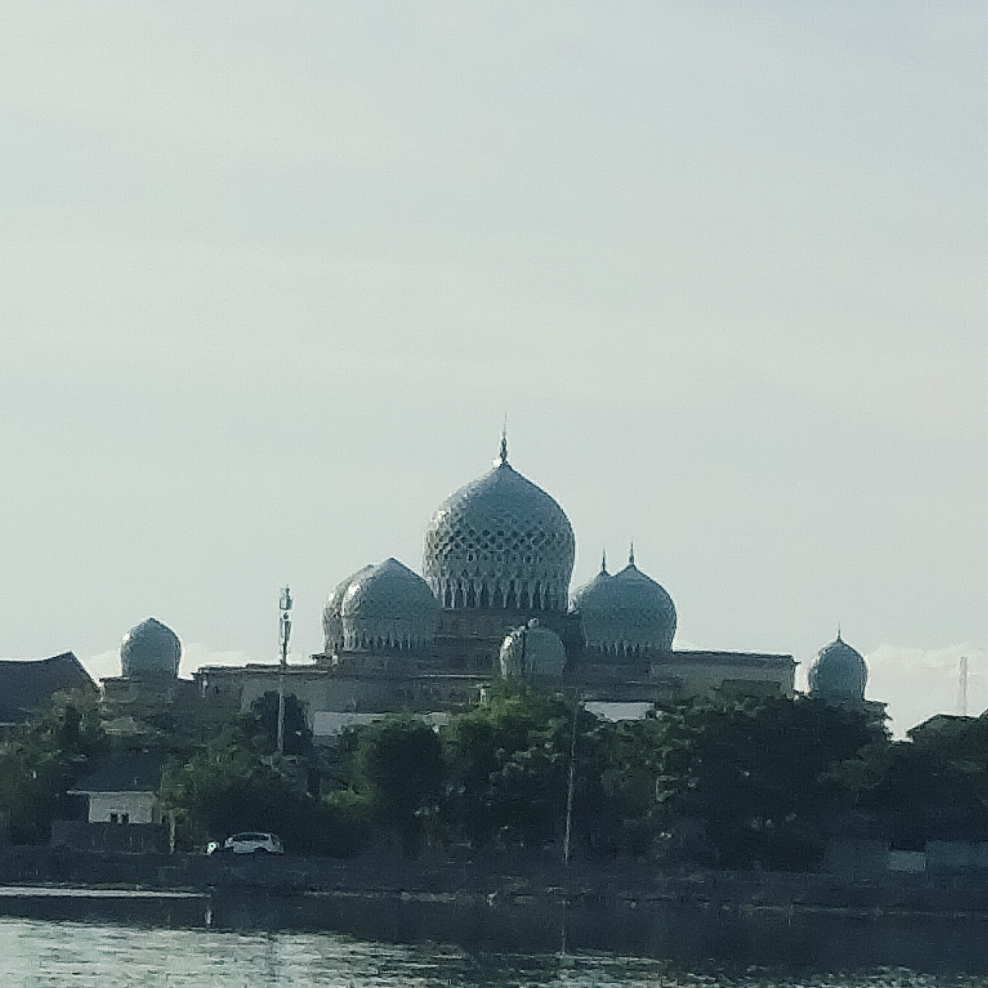 The image depicts a mosque with a central, intricately patterned dome and several smaller domes surrounding it. The mosque is located in Lhokseumawe, Indonesia. The building is partially obscured by trees and vegetation, and there is water in the foreground. A communications tower is visible near the mosque. A building and a white car are visible near the trees. The sky is overcast.
