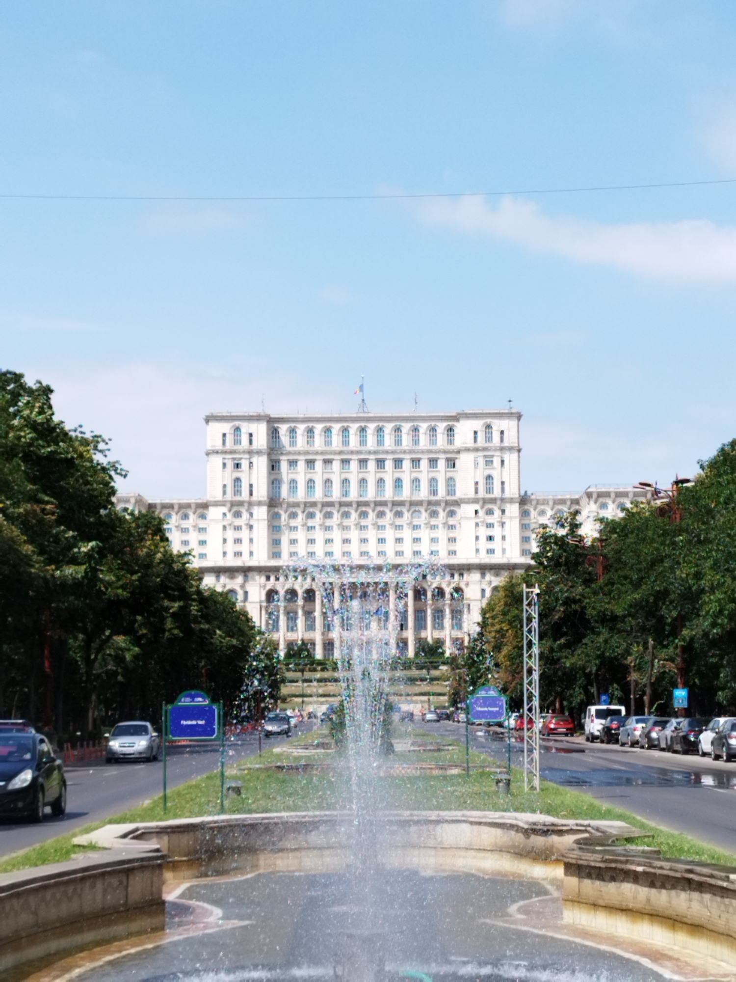 The image depicts a wide avenue in Bucharest, Romania, leading towards the imposing Palace of the Parliament. A large, white, multi-story building with numerous windows and architectural details dominates the background. In the foreground, a fountain sprays water, with its mist partially obscuring the view of the street and surrounding trees. Cars are visible on the road, some parked and others in motion. Blue street signs with text are placed along the avenue. The sky is a clear, light blue with a few wispy clouds. Lush green trees line the sides of the avenue.