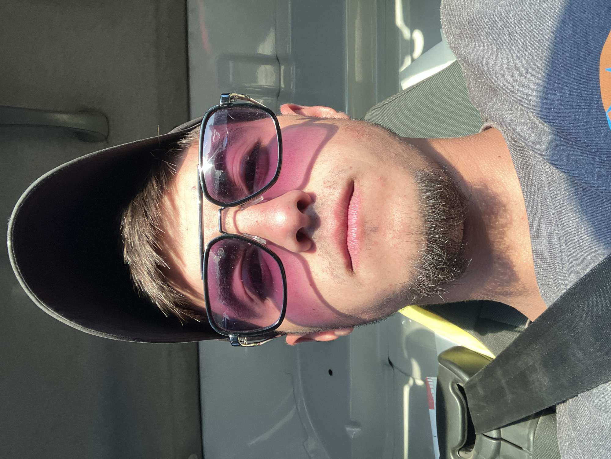 A medium shot depicts a person wearing a gray t-shirt, black cap, and sunglasses with tinted lenses. The person is inside a vehicle, as indicated by the visible seatbelt. The vehicle's interior appears to be white. The person's facial hair is light brown. The location cannot be determined from the image.