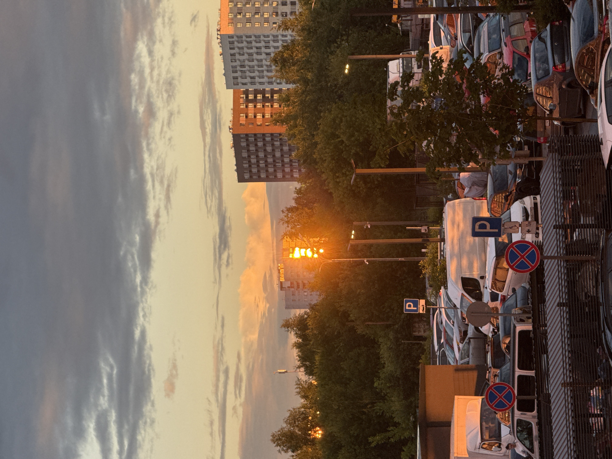 This is a vertically oriented photograph of a street scene in Drozhzhino, Russia, at dusk. The sun is low in the sky, casting a warm, golden light that illuminates the clouds and the windows of a multi-story building. The sky is filled with a mix of gray and pale yellow clouds, suggesting a transition from day to night.
In the foreground, a row of parked cars and vans line the street. Several of these vehicles are white vans, and others are passenger cars in various colors. Visible parking signs with the letter "P" are posted, indicating parking areas. Two circular red and blue traffic signs, likely indicating a prohibition, are also present. The vehicles are parked alongside a metal fence and some trees.
Behind the parked vehicles, dense green trees form a line along the street. Beyond the trees, several tall residential buildings are visible. One building is a striking orange color, contrasting with the gray and white facades of the buildings next to it. The windows of these buildings reflect the warm light of the setting sun.
Streetlights are also visible, some illuminated, contributing to the overall ambiance of dusk. The overall mood of the image is peaceful and serene, capturing the quiet beauty of a suburban street at the end of the day. No people are clearly visible in the frame.