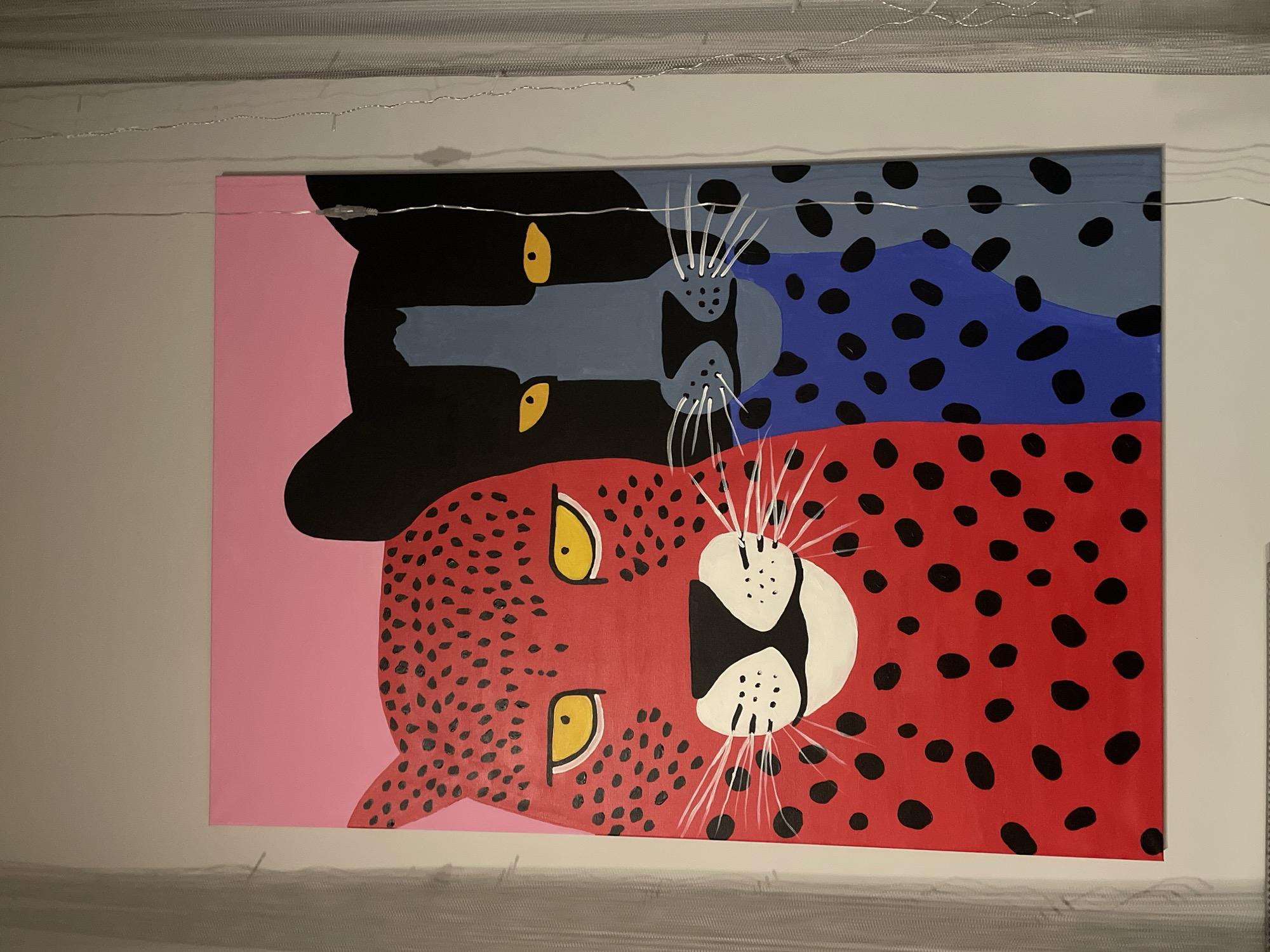 A photograph shows a large rectangular painting hanging on a wall. The painting features three vertically stacked stylized depictions of leopards or similar spotted felines. The top feline is primarily black with a blue-grey muzzle and bluish-purple spots. The middle feline has a similar muzzle with a black head, while the lower feline is red with black spots, and a white muzzle with black markings. Each feline has yellow eyes and white whiskers. The background to the left of the felines is pink. The painting is lit by string lights above and below, and the wall on which the painting hangs is off-white. The setting appears to be indoors. The painting is not demonstrably connected to Krasnoyarsk, Russia.