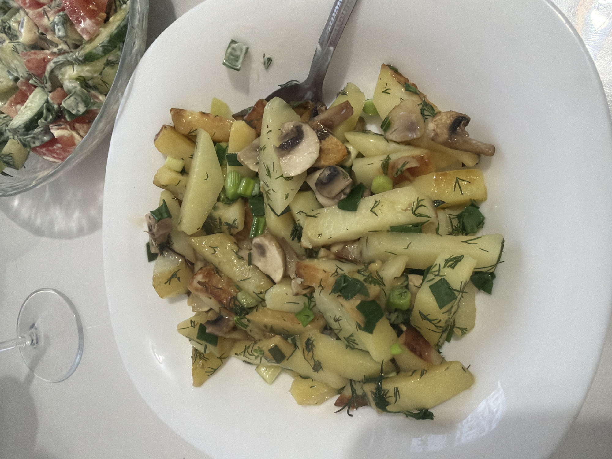 The image shows a white plate with a serving of food. The main component of the dish appears to be potato pieces, some of which are slightly browned. Sliced mushrooms and green onions are mixed in with the potatoes. A silver spoon rests on top of the food. Beside the plate, on the left, is a glass bowl containing what seems to be a salad composed of cucumbers, tomatoes, and a creamy dressing. A wine glass stem is also visible on the left side of the frame. The image doesn't provide enough information to determine the specific location or to identify any people, actions, or interactions.