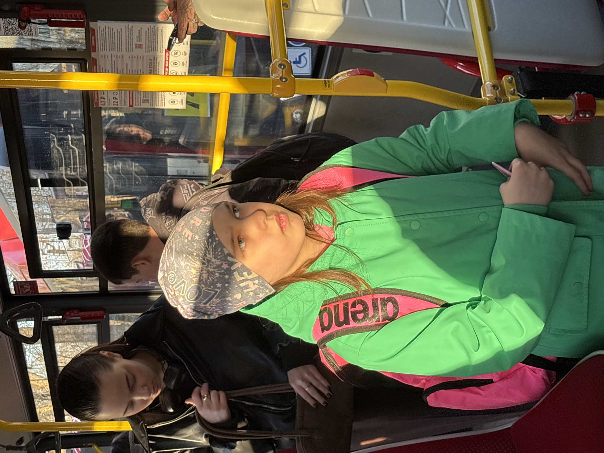 The image depicts a scene inside a public transportation vehicle, possibly a bus, in Perm, Russia. Three people are visible. In the foreground, a young woman with fair skin and light brown hair is wearing a green jacket with a pink "arena" logo, along with a headscarf. She appears to be holding a pen and looking towards the camera. Behind her, two other passengers are visible. One person is wearing a grey hat with text "LOVE" and the other has black hair and is wearing headphones. The bus interior features yellow handrails and advertisements. The vehicle appears to be moving, as suggested by the blurred scenery visible through the windows.