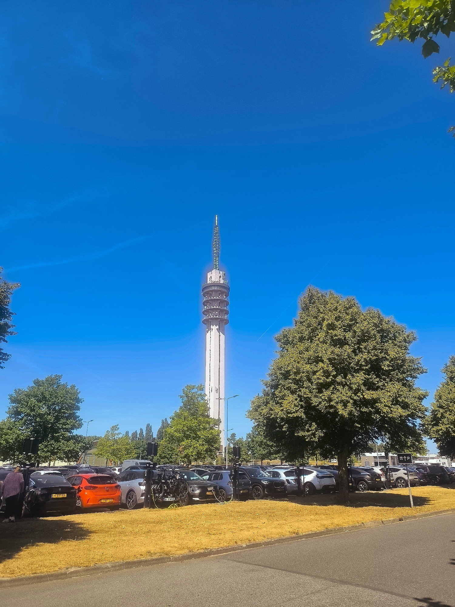 The image depicts a clear, bright day with a vibrant blue sky. In the background, a tall, slender white tower with multiple levels and antenna at the top dominates the skyline. In the foreground, a parking lot is filled with various cars, some of which are electric vehicles plugged into charging stations. Lush green trees are scattered throughout the scene, with one large, prominent tree in the middle right of the frame. A patch of dry, yellow grass runs along the edge of the parking lot, bordering an asphalt road. Two individuals, one person in a dark purple top and another in a dark top and light-colored pants, are visible on the left side of the image, near the parked cars. The overall scene suggests an outdoor environment, likely a public space with facilities for parking and charging electric vehicles, set against the backdrop of a significant urban landmark, possibly the TV tower of Lelystad, Netherlands.