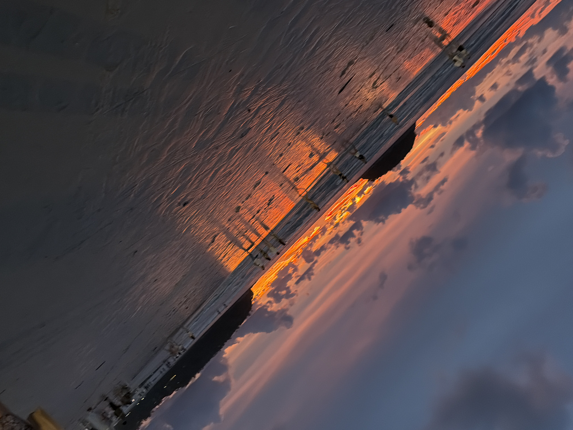 The image depicts a beach scene at sunset. The sand, textured with ripples, occupies the foreground and midground, reflecting the vibrant hues of the setting sun. The sun's light casts long shadows from unseen objects, possibly beach chairs or umbrellas, across the sand. A dark, silhouetted structure is visible in the distance, partially obscured by the sand. The horizon line is dominated by a band of intense orange and yellow light from the sun, transitioning into soft pinks and purples in the sky above. Darker, wispy clouds are scattered across the sky, contrasting with the lighter, illuminated clouds. The overall mood is serene and peaceful, characteristic of a sunset. The scene appears to be set in Karon, Thailand, indicated by the tropical aesthetic suggested by the beach and the time of day, which is late afternoon to early evening. There is no visible text in the image.