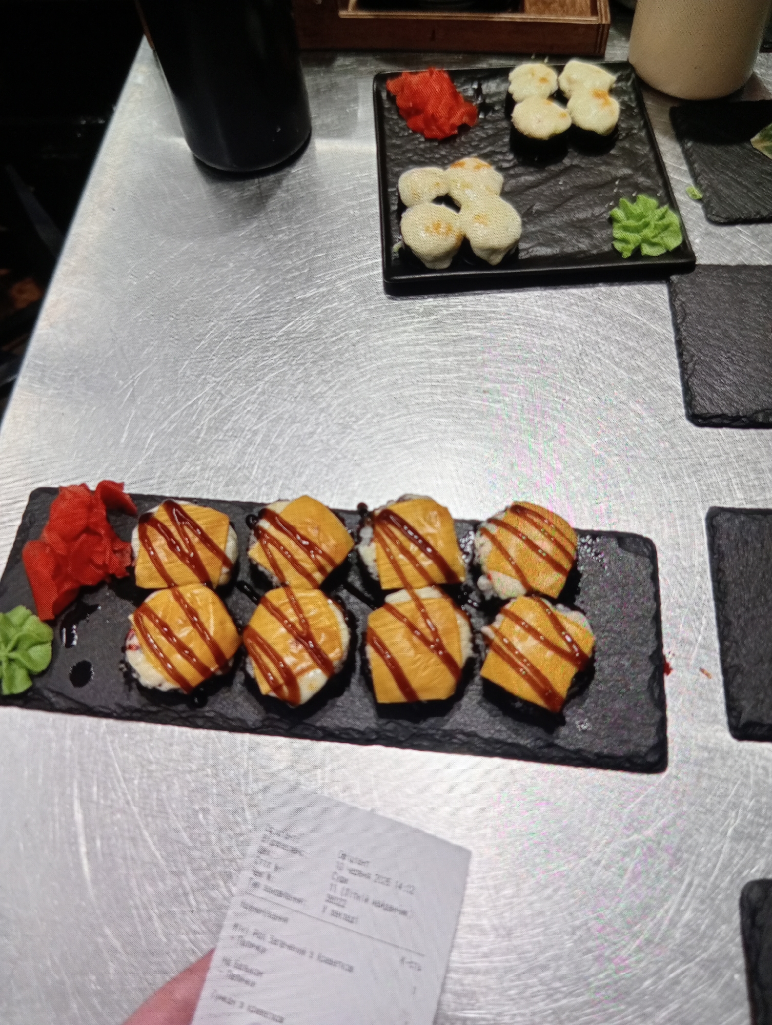 A close-up, eye-level shot captures a table setting with sushi and a receipt. The table has a metallic, brushed surface. In the foreground, a black rectangular plate holds eight pieces of sushi, each topped with a yellow layer and drizzled with a dark sauce. To the left of the sushi are two small piles of bright red pickled ginger and a small mound of green wasabi. In the background, another black square plate holds four pieces of what appear to be baked scallops or another type of seafood, also with a creamy topping. Adjacent to this plate is a small serving of green wasabi. Further in the background, a dark bottle and a light-colored ceramic mug are visible. A portion of a printed receipt is visible in the lower right corner of the frame, held by a hand. The overall scene suggests a meal at a restaurant.