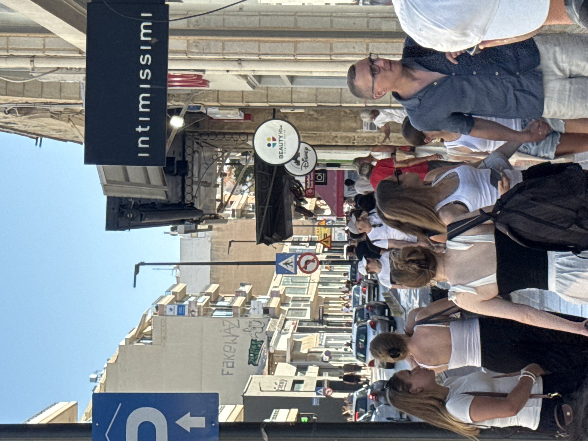 The image depicts a bustling street scene in Chania, Greece. In the foreground, a man wearing glasses and a blue patterned shirt holds hands with a woman in a white tank top and denim shorts. Behind them, other pedestrians navigate the crowded sidewalk. The background features a row of shops, including "intimissimi" and "BEAUTY Yu." Cars are visible on the street, and buildings line both sides of the road. The sky is clear and blue.