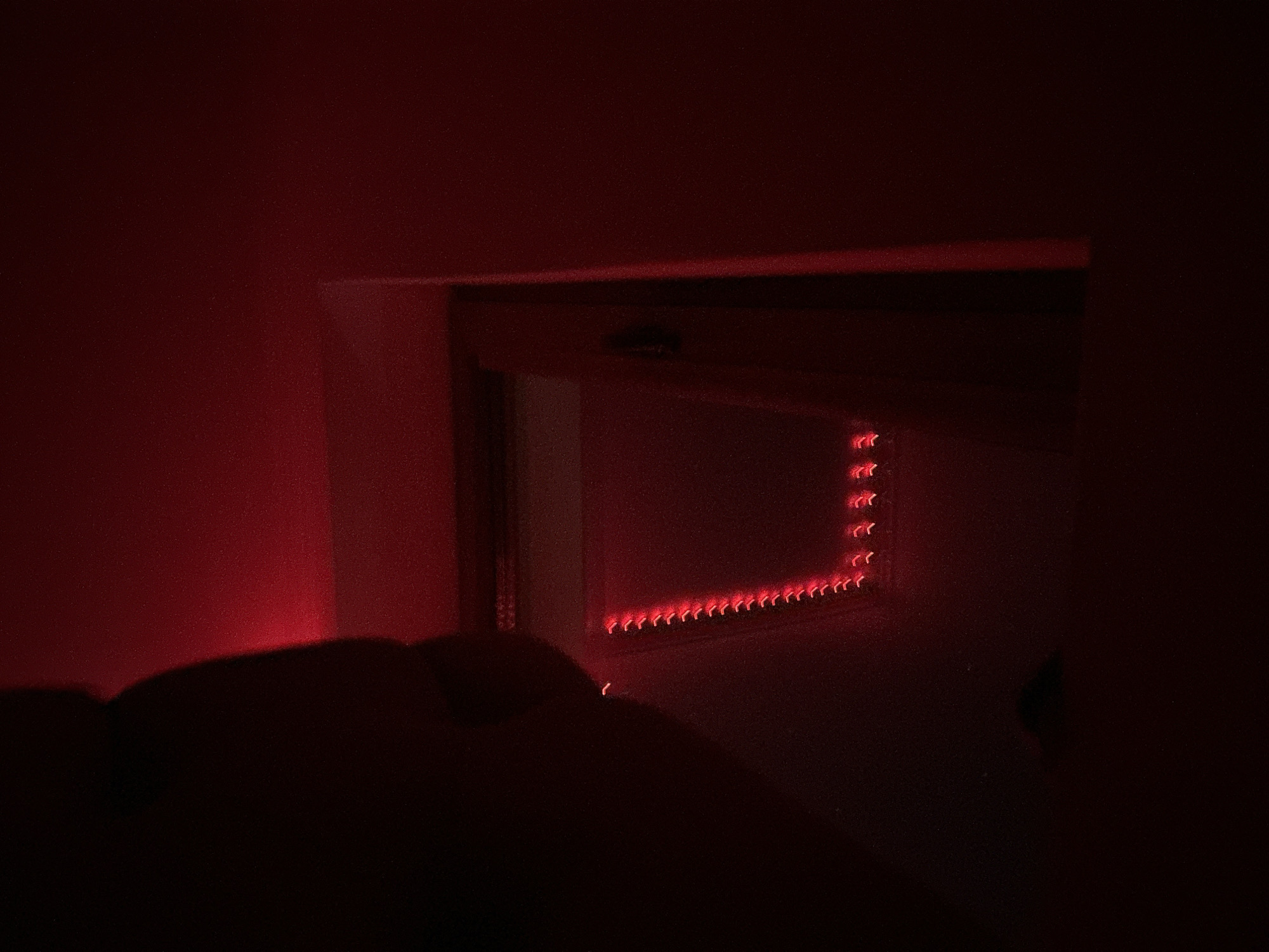 The image depicts a dimly lit room with red ambient lighting. On the left side, a dark silhouette of a person's head and shoulder is visible, partially obscuring the background. The background shows a wall with a window or opening where a string of red LED lights is installed, forming a curved pattern. The lights create a bokeh effect. The environment suggests an indoor setting, possibly a bedroom or entertainment space. There is no information in the image to indicate the location in the city of Shumen, Bulgaria.