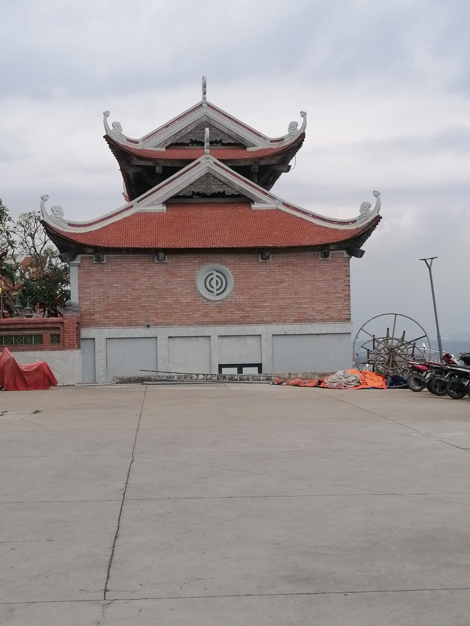 This image captures a two-story building with traditional East Asian architectural elements, resembling a temple or pagoda, under a uniformly grey, overcast sky in Hong Gai Ward, Vietnam. The building features a reddish-brown brick facade on its lower level, centered with a distinctive circular decorative window displaying a geometric pattern. Its most notable architectural details are the tiered, reddish-brown tiled roofs with intricately curved white eaves that sweep upwards, adorned with ornate finials at the corners.
The ground floor of the building presents a more utilitarian appearance with multiple grey, closed panels resembling garage doors, and a smaller access door to the left. In the foreground, a wide, flat concrete plaza or courtyard extends towards the viewer, marked by expansion joints.
Various objects are scattered around the courtyard, suggesting ongoing activity or storage. On the left, a large red tarp covers an unseen object. A ladder is propped near the building's entrance. To the right, several motorcycles are parked in a row, and a prominent large wooden spoked wheel, possibly an old cart wheel or a decorative item, rests on the ground amidst other bundled materials and tarps. A tall, modern street lamp stands to the right of the building. The scene is devoid of people, and the cloudy sky suggests a subdued daylight. No visible text is present.