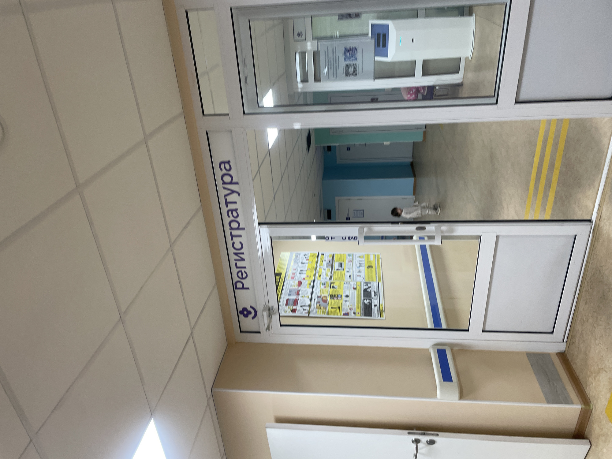 The image shows the entrance to a registration area, identified by the sign "Регистратура" (Registration) in Russian, above a glass door. A small child in a light-colored outfit is visible through the doorway, standing on a polished floor marked with yellow lines. Inside the registration area, there is a white kiosk-like machine with a screen and a slot, and a poster is displayed on a wall. The hallway outside the registration area features a light-colored wall with a blue and white handrail. To the left of the registration entrance, a white door with a silver handle is partially visible. The ceiling is a standard white tiled grid. The scene appears to be an interior hallway of a medical facility or a public service building.