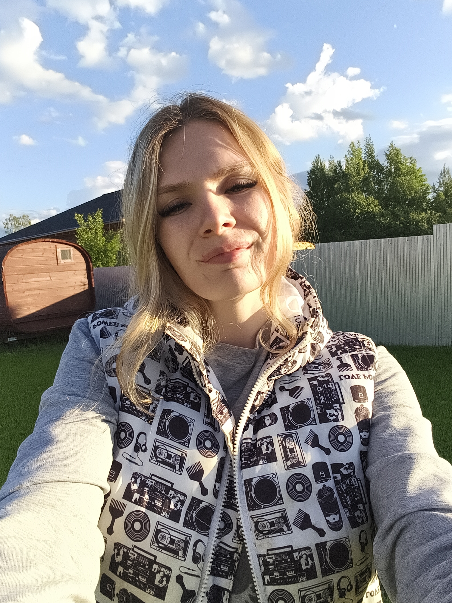 A woman is taking a selfie outside on a sunny day. She has long blonde hair, and is wearing a gray long-sleeved shirt with a sleeveless jacket with black and white designs. Behind her is a green lawn, a metal fence, some trees, and a small wooden building. The location may be in the city of Русский Вожой, Russia.