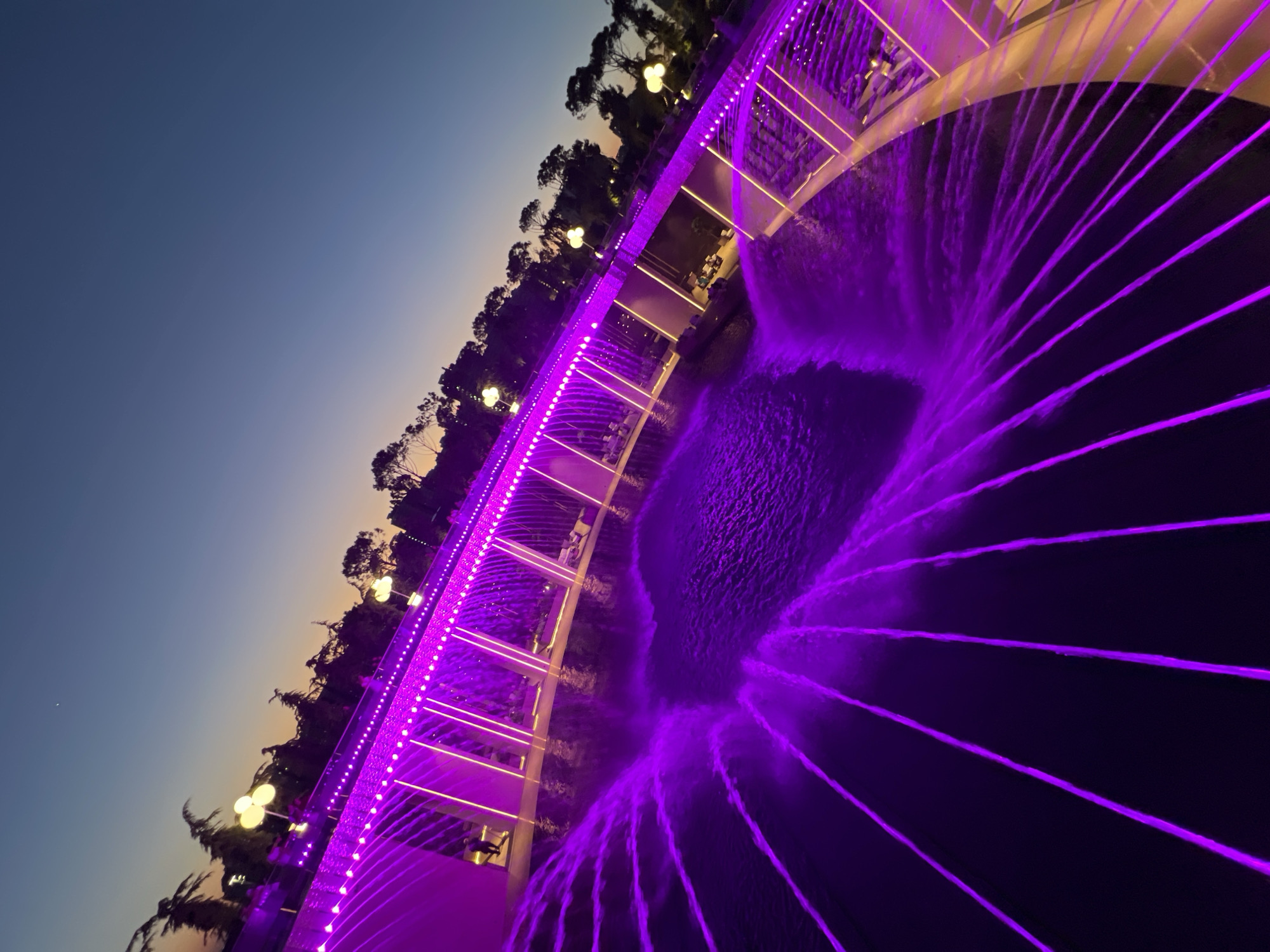 The image depicts a contemporary water fountain feature illuminated with vibrant purple lights at dusk. The fountain is situated in a park or public space, characterized by lush trees silhouetted against a gradient sky transitioning from a warm orange near the horizon to a deep blue overhead. The fountain itself features multiple jets of water, which are also lit with the same purple hue, creating a dynamic and visually striking display. A modern, possibly a bridge or elevated walkway, with minimalist design and integrated lighting, spans across the fountain area. Several figures, appearing as silhouettes, can be seen on and around the walkway, suggesting public use of the space. The overall impression is one of a well-maintained, aesthetically pleasing urban recreational area designed for evening enjoyment. This fountain is located in the City of Krasnodar, Russia, specifically in the "Park of Memory" or "Park of the First Cosmonaut" on the embankment of the Kuban River.