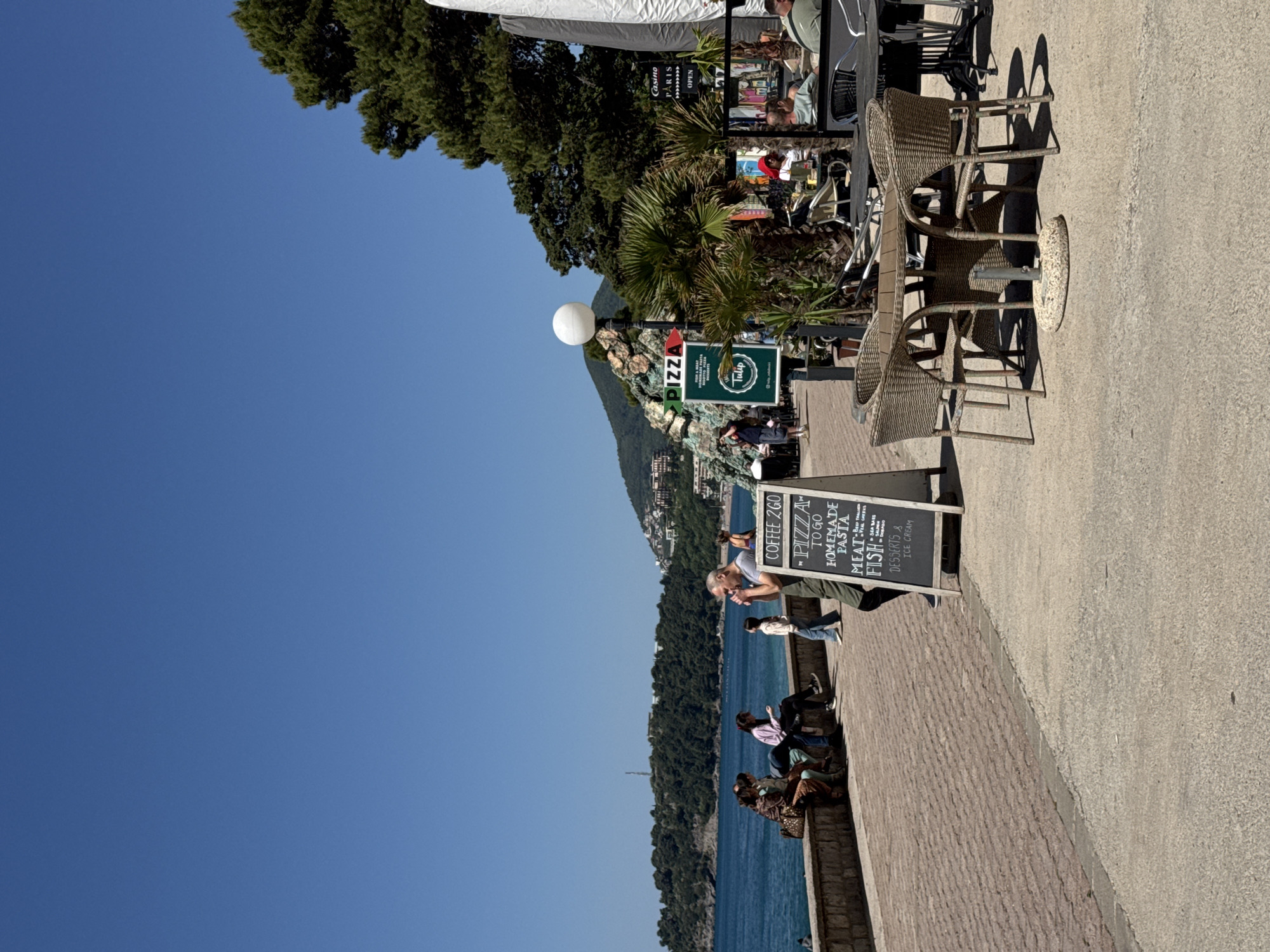 This outdoor photograph captures a sunny seaside promenade in Rafailovići, Montenegro, bustling with activity. The scene is dominated by a clear blue sky and calm, deep blue waters of the Adriatic Sea on the left. Across the water, lush green, tree-covered hills rise, with some distant structures nestled among them.
On the right side of the image, a paved promenade, comprising sections of light concrete and reddish bricks, hosts an outdoor cafe or restaurant area. Several wicker chairs and tables are arranged under white awnings, with some patrons seated and engaged in conversation or looking at menus. Green palm trees and other foliage line the promenade, adding to the Mediterranean feel.
Various signs are visible: a prominent black sandwich board advertises "COFFEE 2GO," "PIZZA TO GO," "HOMEMADE PASTA," "MEAT," "FISH" (including "SEA BARS," "SHRIMP," "SQUID," and "OCTOPUS"), "DESSERTS & ICE CREAM." Nearby, a sign with an arrow points to "PIZZA," and another reads "Tulip." Further back, a "Casino PARIS OPEN" sign is discernible. A spherical white street lamp stands out against the green hills.
Several individuals are present: some are seated at the cafe, while others stroll along the promenade or sit on the low wall overlooking the sea. One man in a light blue shirt stands near the sandwich board, gazing towards the water. The strong shadows cast by the furniture indicate it's daytime, likely mid-morning or early afternoon, under clear, bright weather conditions. The overall atmosphere is relaxed and leisure-oriented, typical of a coastal resort town.
