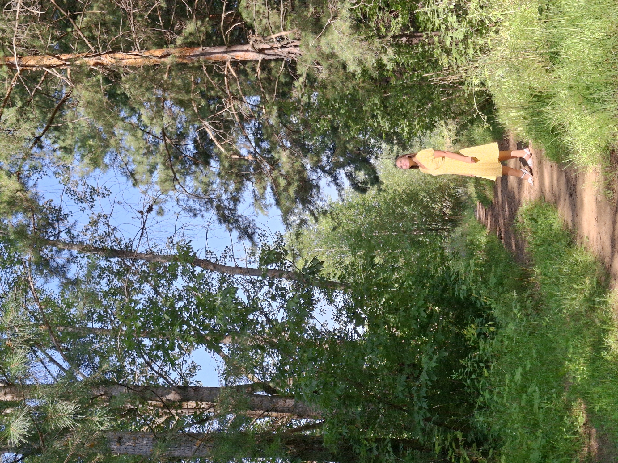 A young woman in a yellow dress stands on a dirt path in a forest. She is looking up at the sky. The path is lined with trees and green foliage. The sky is blue with some clouds. The woman is wearing white sandals.