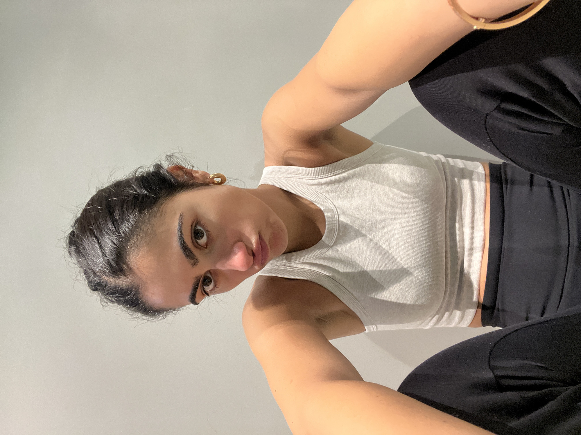 A woman with dark hair pulled back from her face, wearing a light gray tank top and black pants, is sitting with her knees up, looking at the camera. She is wearing a gold earring and a gold bracelet. The background is a plain, light gray wall. There is no indication of the location being Paris, France.