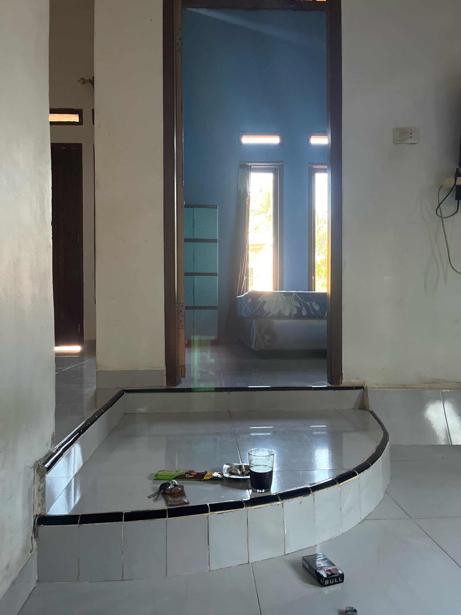 This is an eye-level, interior shot of a house in Kebumen, Indonesia. The image captures a view through an open doorway into another room. In the foreground, a semi-circular tiled platform with a black trim is visible. On this platform, a glass of dark liquid, a packet of cigarettes labeled "BULL," a set of keys with a leather fob, and a small plate with what appears to be food are placed. The floor in the foreground is tiled.
Through the doorway, the adjacent room has blue walls and two windows. Sunlight streams in from the windows, illuminating a bed with a floral patterned comforter. To the left, in the background, a blue cabinet with multiple shelves is visible. The doorway frame is dark brown wood. To the right of the doorway, a white wall features a light switch. The lighting suggests it is daytime. The overall scene depicts a domestic interior, possibly a living area or hallway leading into a bedroom.