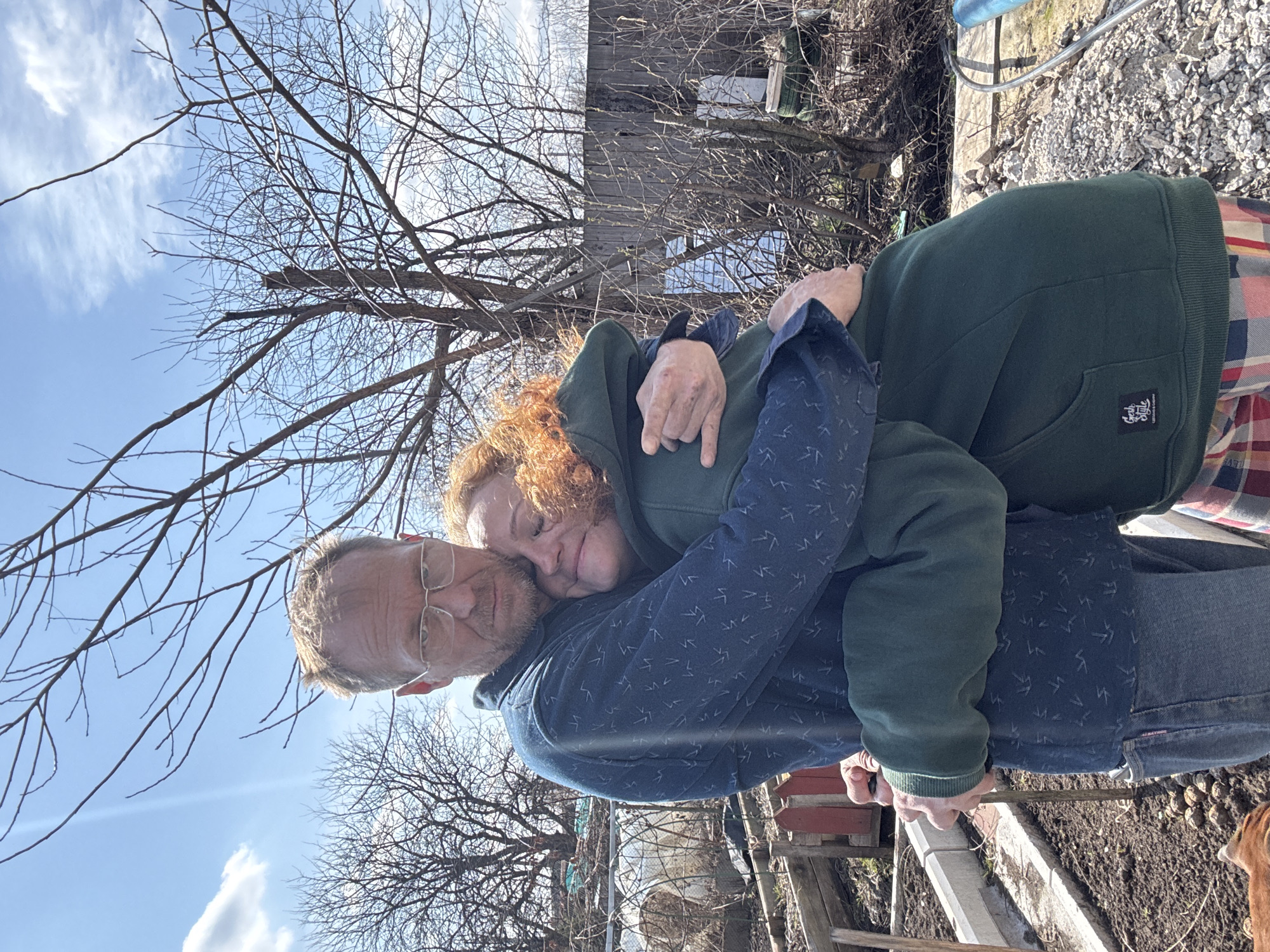 An affectionate moment between a man and a woman is captured in this outdoor vertical shot, likely in a backyard or garden in Chaikovsky, Russia. The image is rotated 90 degrees counter-clockwise from its original capture.
The man, with reddish-blonde hair and a beard, wears glasses and a dark blue patterned hoodie over a lighter blue shirt and blue jeans. He is embracing the woman, holding a small dark object in his hands at waist level. The woman, with striking curly red/auburn hair, rests her head tenderly on his shoulder. She wears a dark green hoodie with a front pouch pocket, which bears a small black tag reading "Fun Style" and "Casual Wear" below it. A plaid shirt is visible underneath her hoodie. Both appear comfortable and at ease, conveying a sense of warmth and connection.
The setting is an outdoor space dominated by numerous bare-branched deciduous trees, suggesting the season is late winter or early spring. The sky is bright blue with scattered white clouds, indicating a sunny day. In the background, various elements of a lived-in outdoor area are visible: wooden structures that could be fences or parts of a shed, a metallic or plastic frame that might belong to a greenhouse, cultivated garden beds with concrete borders, and some gravel. A blue pipe and a small, light brown dog are partially visible on the right side of the frame, near the ground.