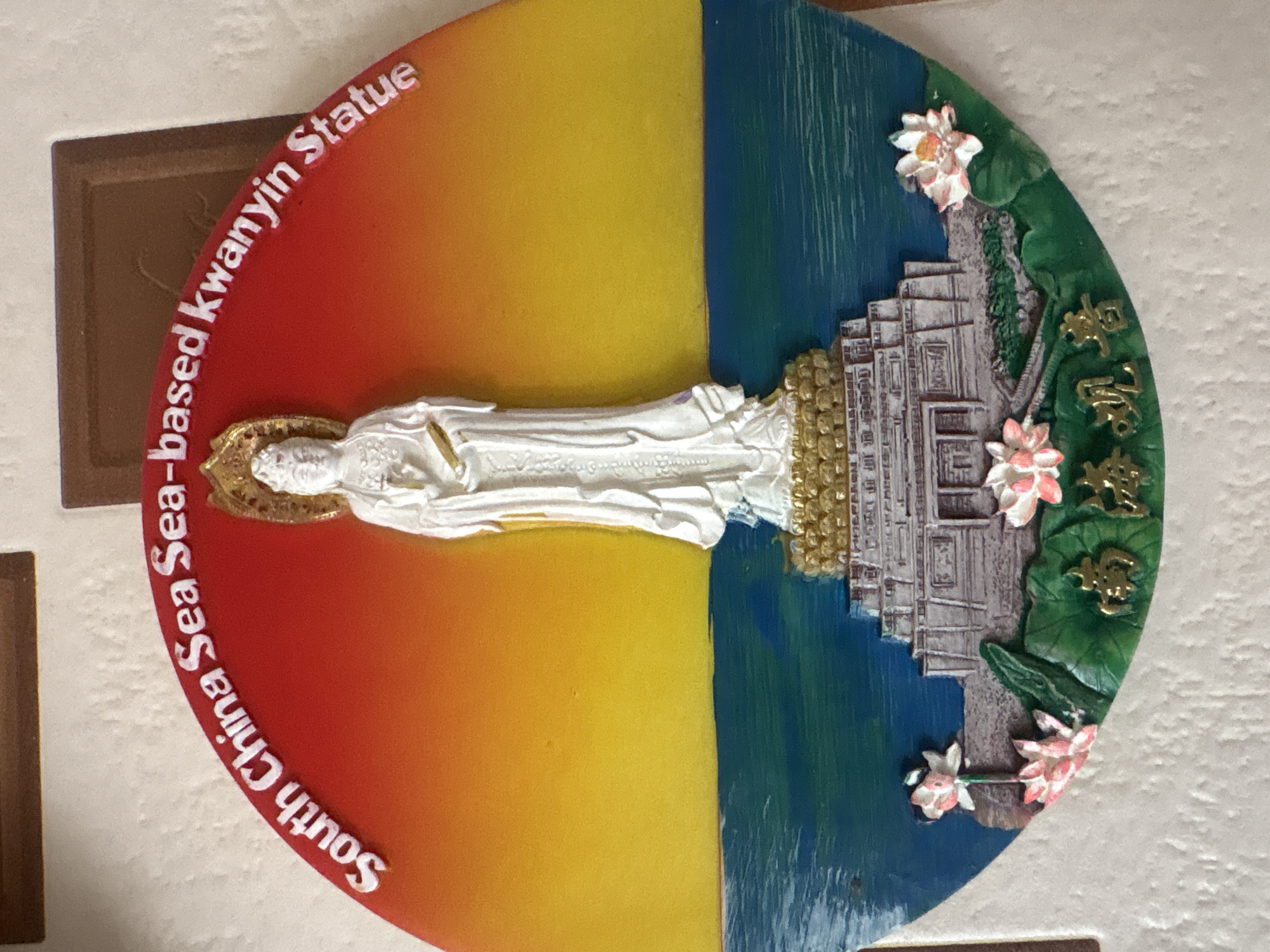 The image shows a circular souvenir magnet depicting the South China Sea-based Kwanyin Statue. The magnet is divided into two halves: the left side is an ombre of red and yellow, and the right side features the statue itself, surrounded by blue, green, and pink elements suggestive of water, foliage, and flowers. The statue is white, and there is gold detailing around its head and base. The text "South China Sea-based Kwanyin Statue" is written in white around the edge of the magnet. The magnet is affixed to a textured off-white surface. There is no evidence to suggest this object is located in Krasnoyarsk, Russia.